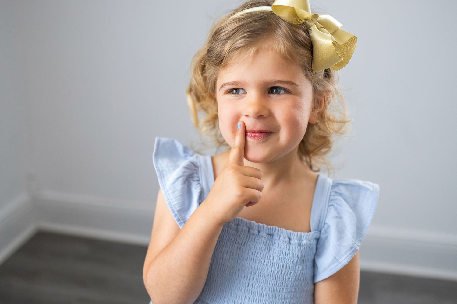 Young child in blue dress with yellow bow poses with finger on chin and playful expression.