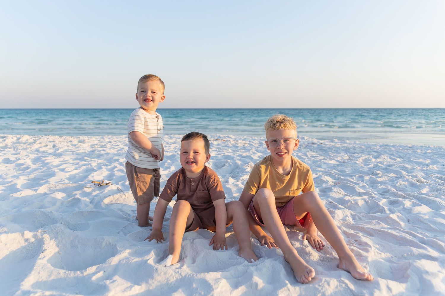 Three children sitting in white sand with ocean waves behind them.