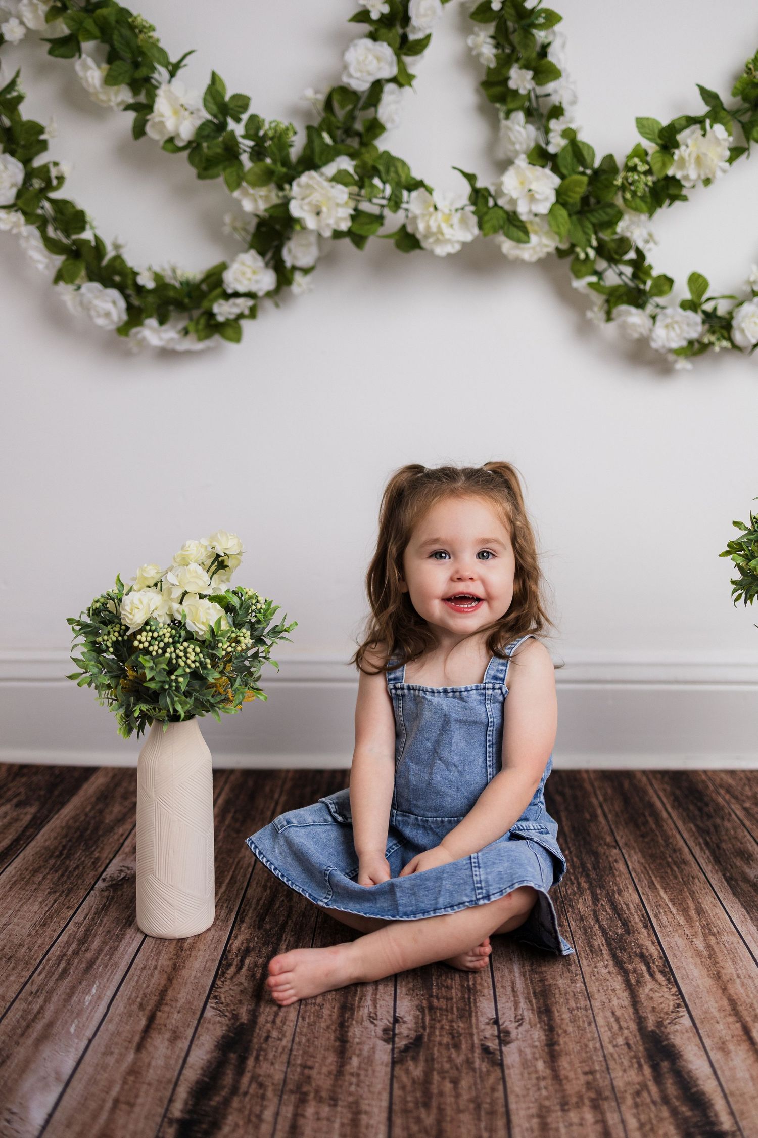 A series of studio photos with floral decor and rustic wood flooring shows a toddler in a denim dress.
