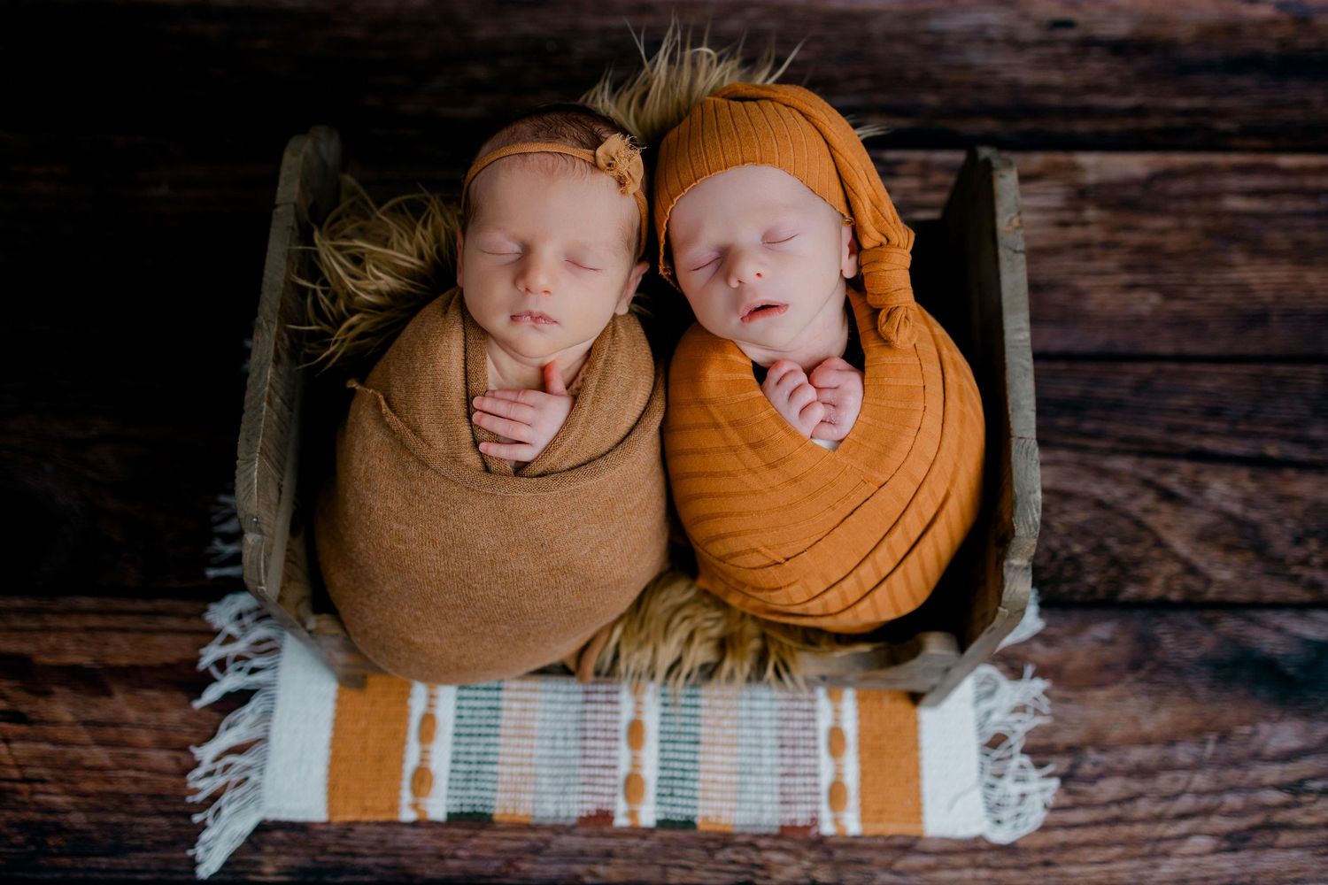 Two swaddled newborns lying side by side in brown and orange wraps on wooden backdrop with striped blanket.