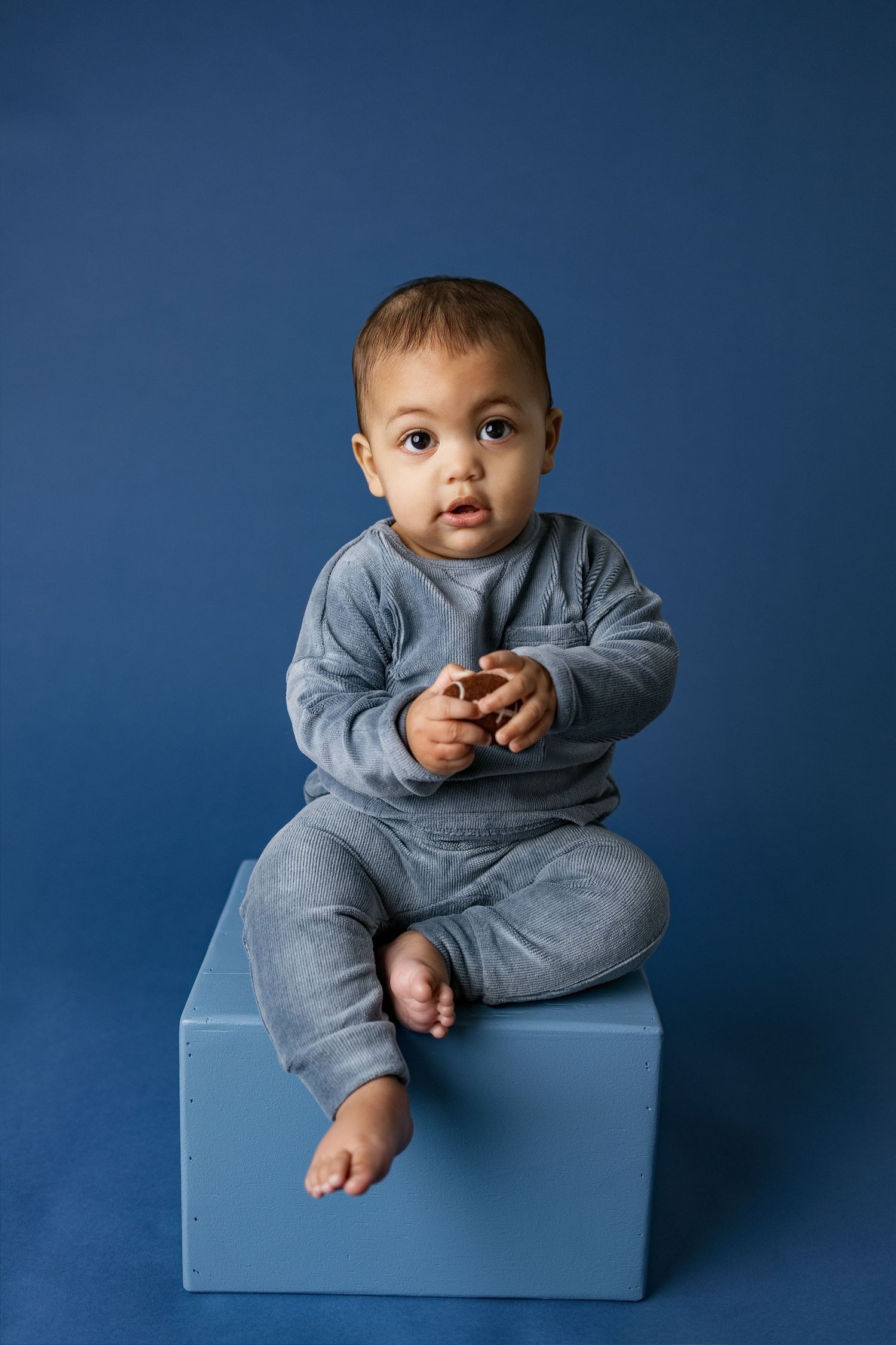 Baby in gray sweatshirt sitting on blue cube against navy background in professional studio portrait series.
