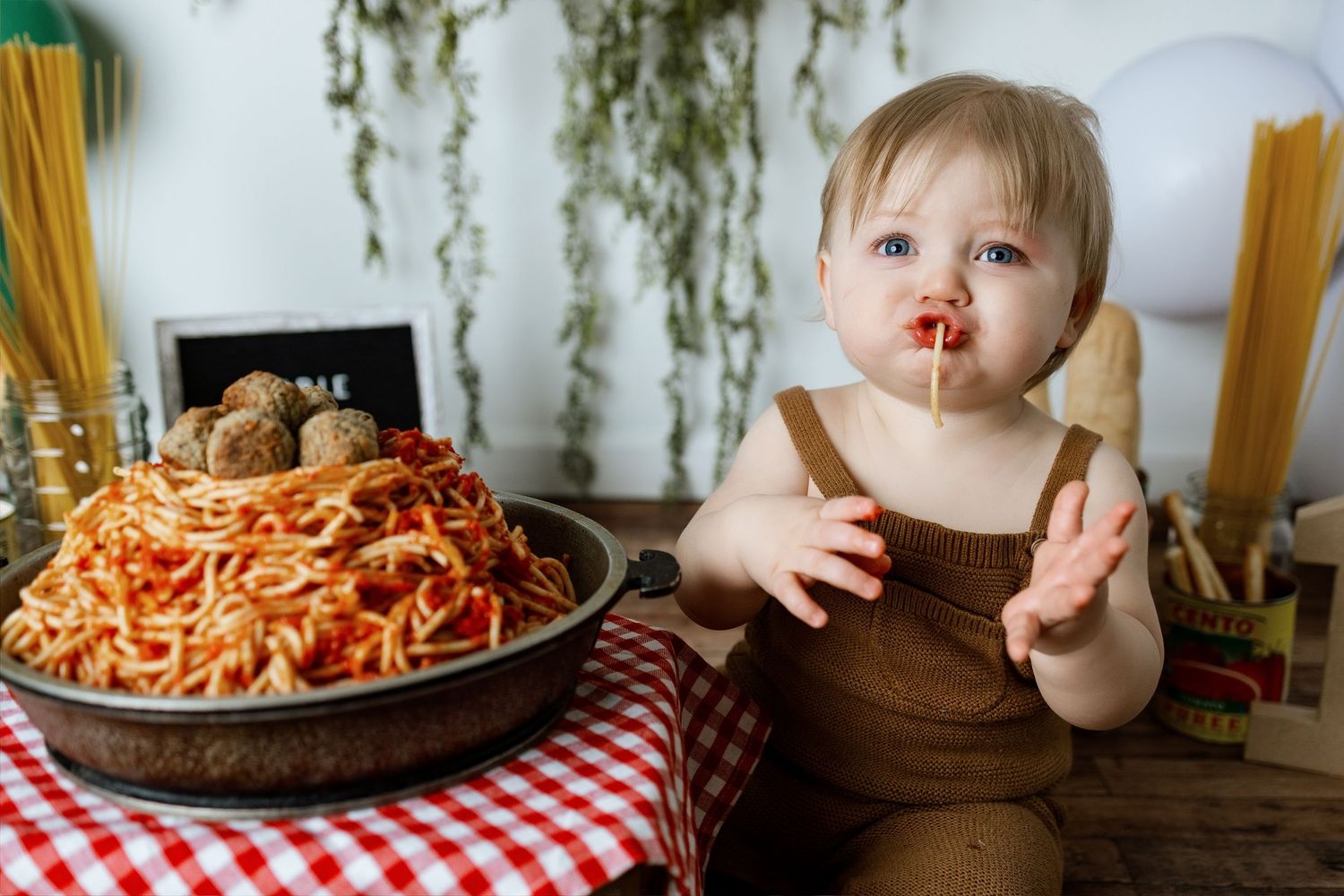 Plates of spaghetti and meatballs on a red checkered tablecloth with fresh herbs hanging above.