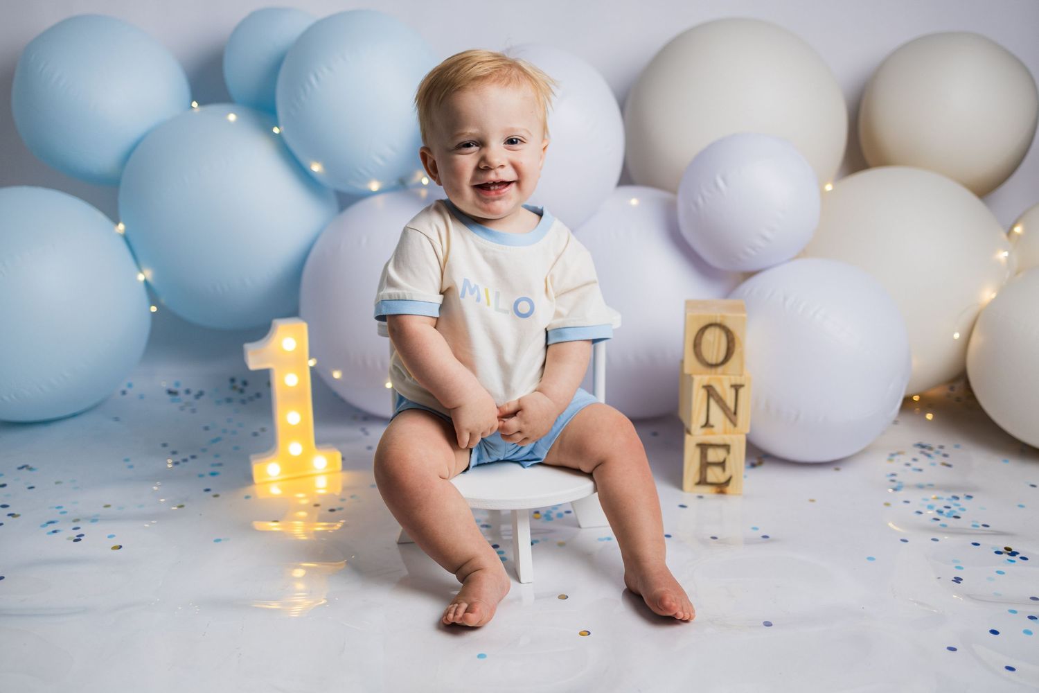 A first birthday celebration setup with glowing number one candle, wooden blocks spelling 'ONE' and pastel balloon decorations.
