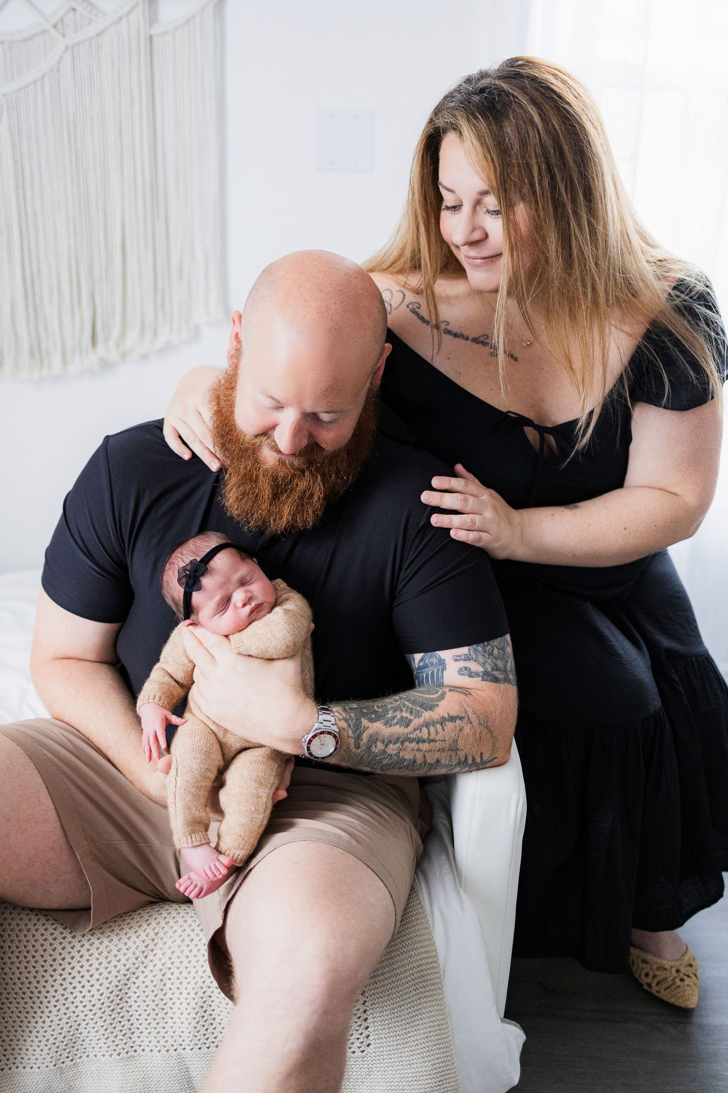 A couple in black clothing shares a tender moment with their newborn baby against a light-colored backdrop.