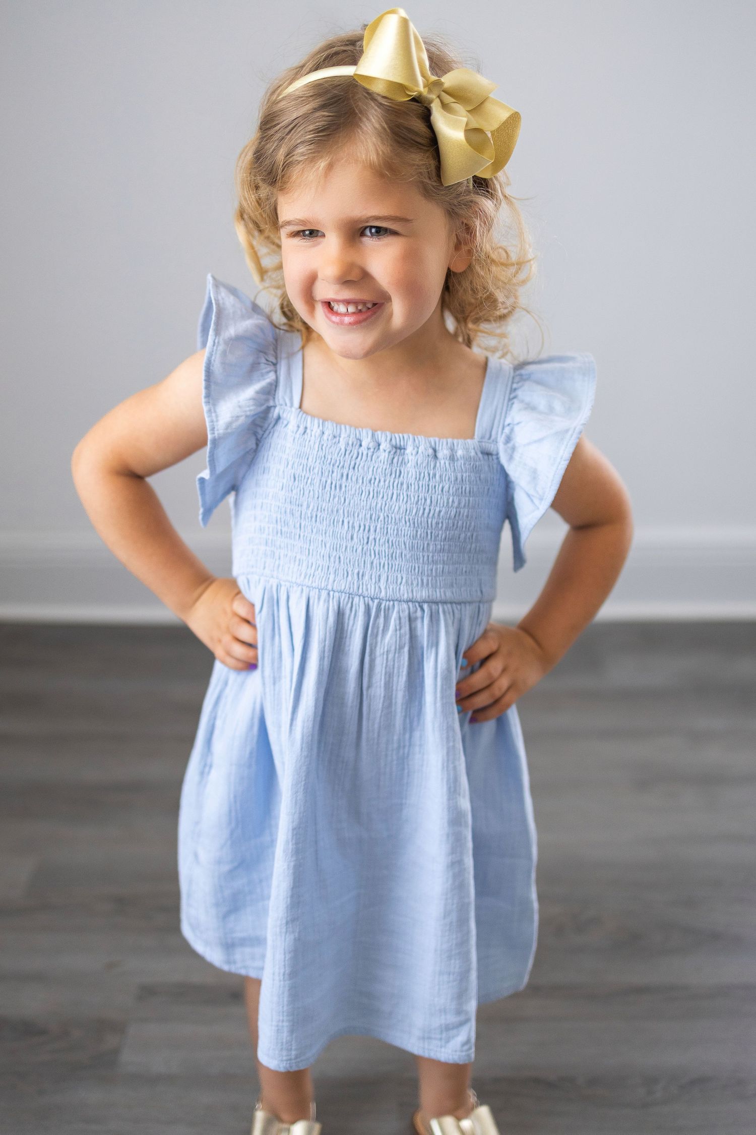 Small child wearing a light blue smocked dress with flutter sleeves and a gold bow hair accessory stands smiling.
