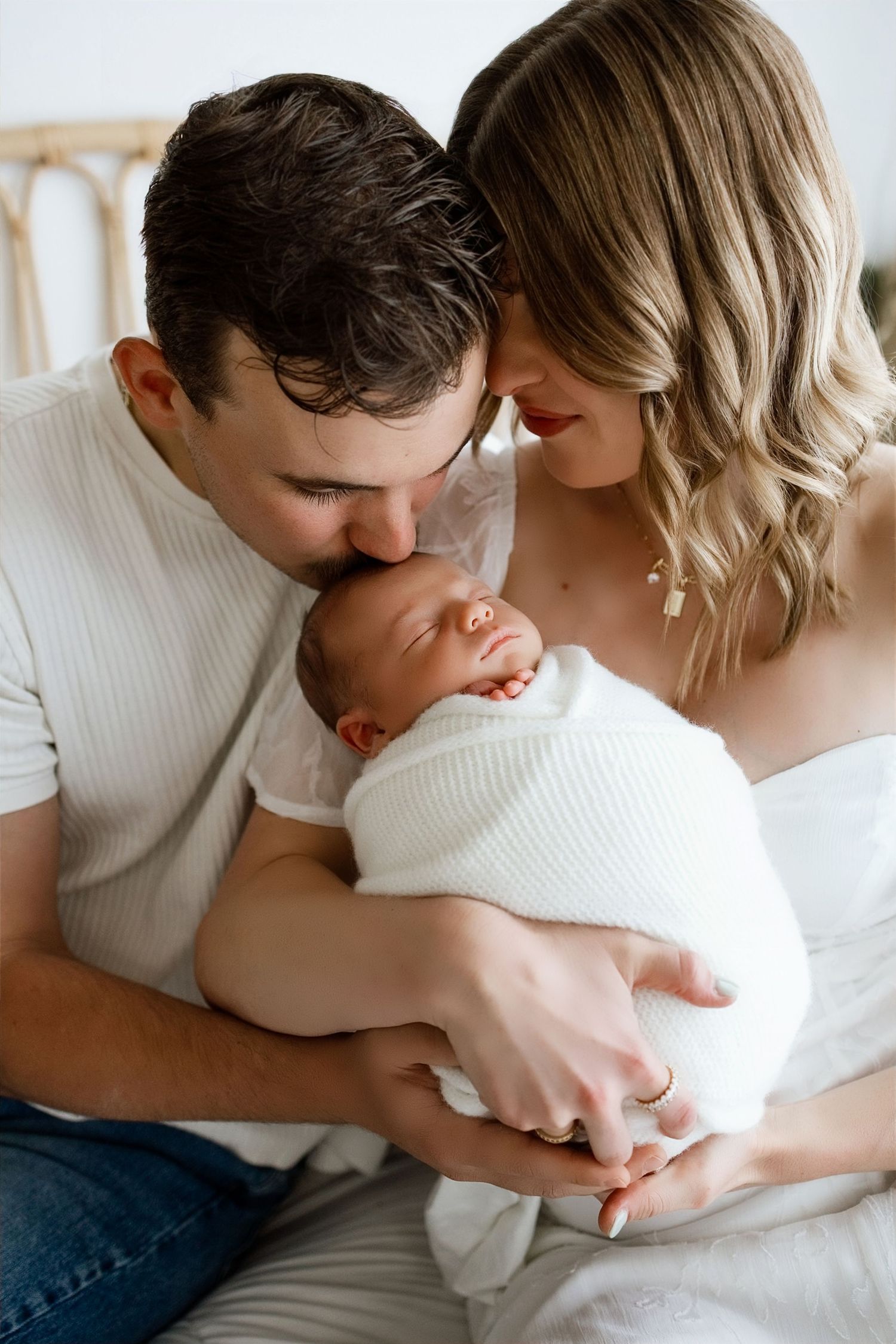 New parents tenderly kiss their swaddled newborn baby in a bright white studio newborn photography session.
