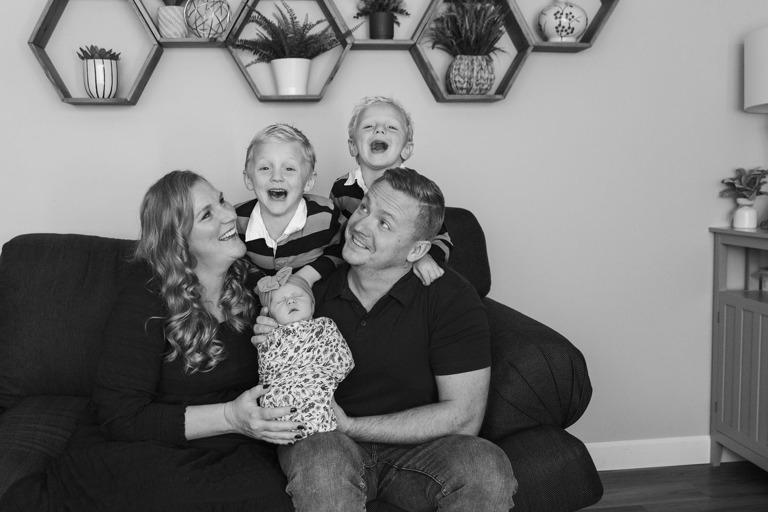 A family laughs together while sitting on a couch with hexagonal wall decorations.