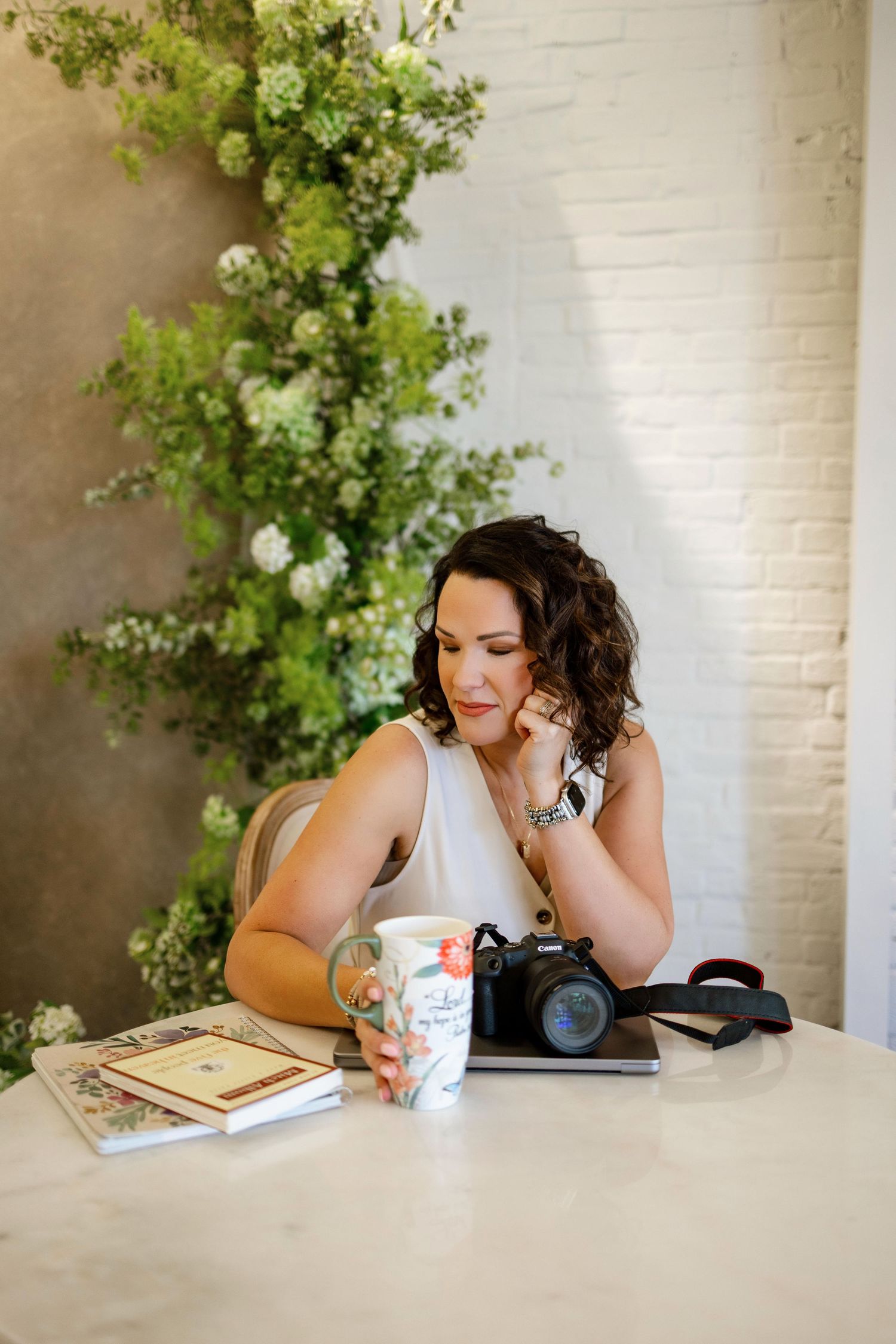 Person seated at table with camera equipment and floral backdrop enjoying coffee and books.