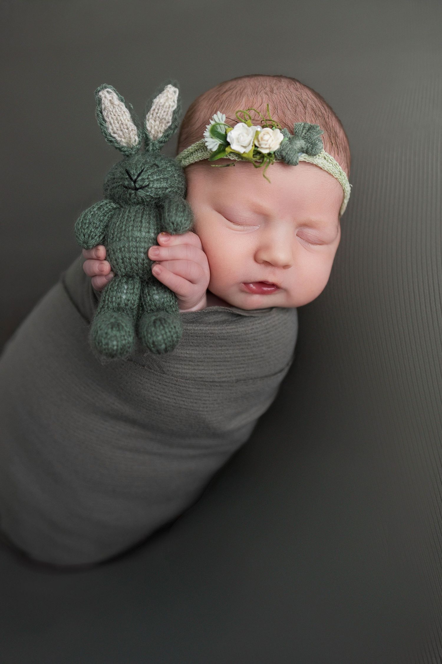 Sleeping newborn swaddled in gray wrap holding knit green bunny toy and wearing delicate floral headband.