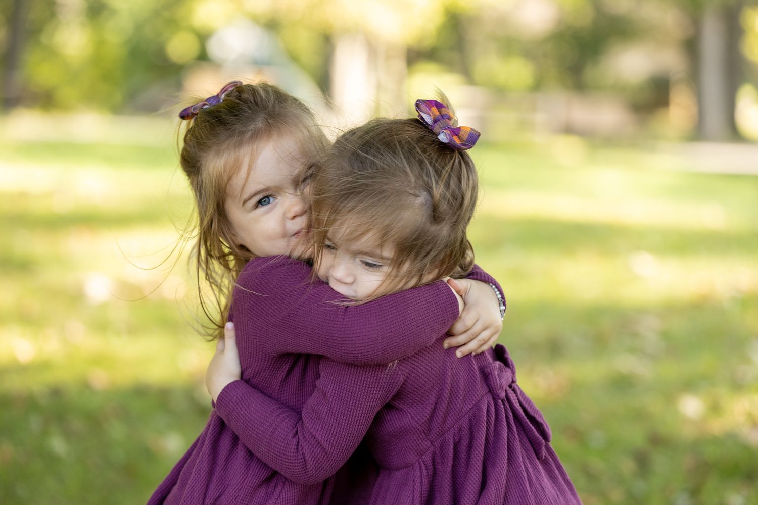 Two young girls in matching purple dresses share a tender hug outdoors in a park setting.