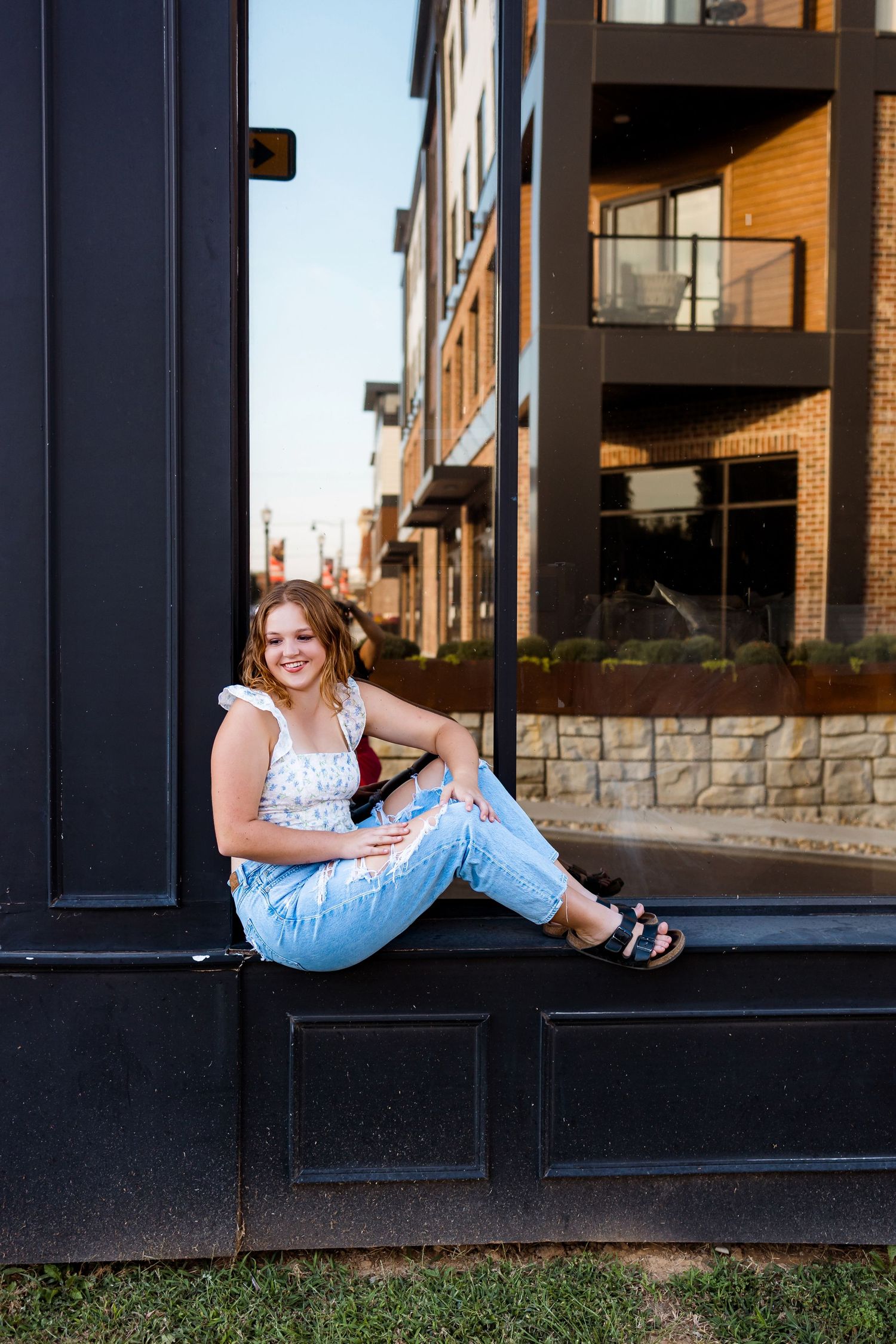 A person in ripped jeans and sandals sits casually on a black windowsill ledge outside a modern apartment building.