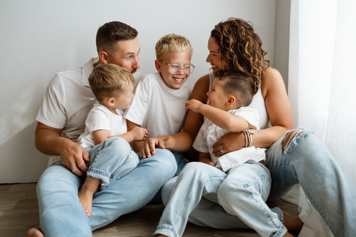 Happy family of five sitting together laughing, parents with three young children in white shirts and jeans.