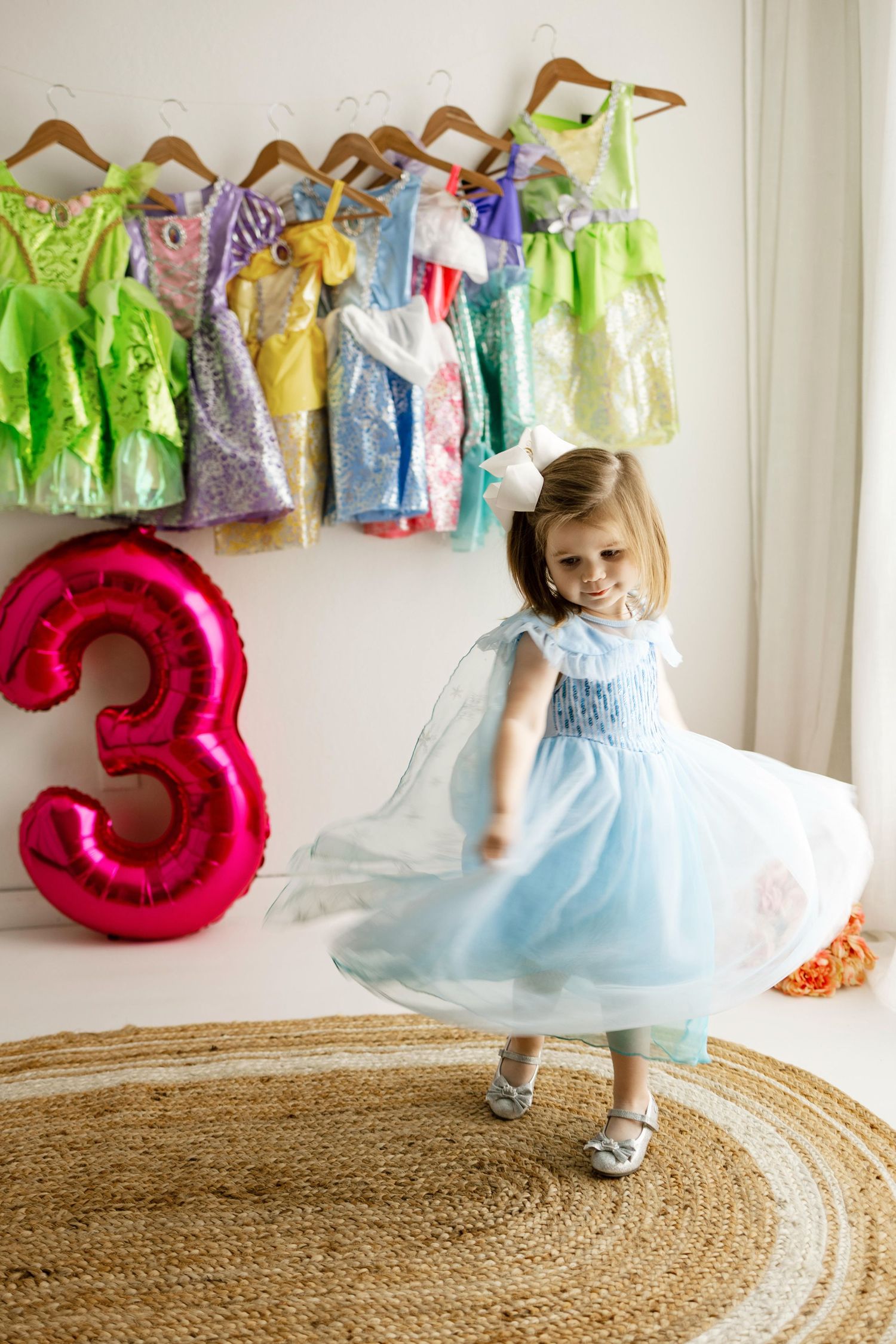 A young child in a light blue dress twirls and spins on a round jute rug with colorful decorations hanging on the wall.