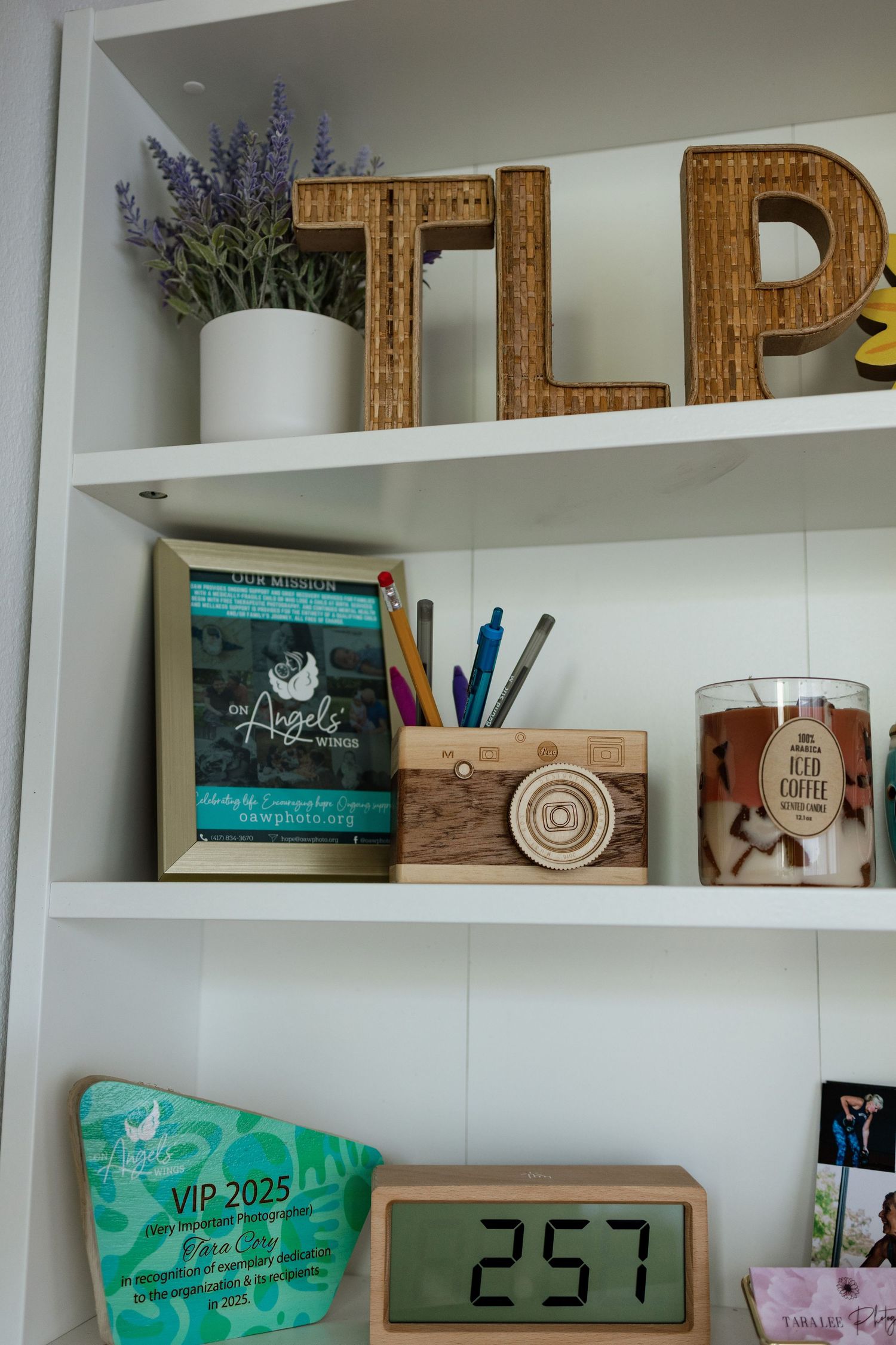 White built-in shelves display wicker TLP letters, lavender plant, wooden camera, pencil holder, iced coffee candle, and VIP 2025 badge.