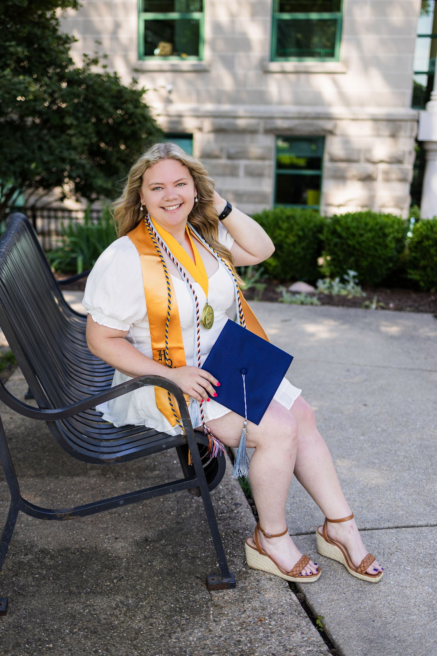 Graduate in white blouse and gold sash sits on outdoor bench holding diploma with academic building behind.