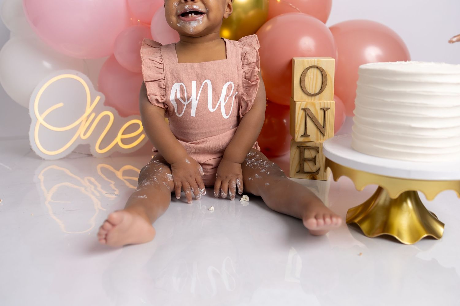 Baby in pink outfit sits by birthday cake with pink balloon backdrop.