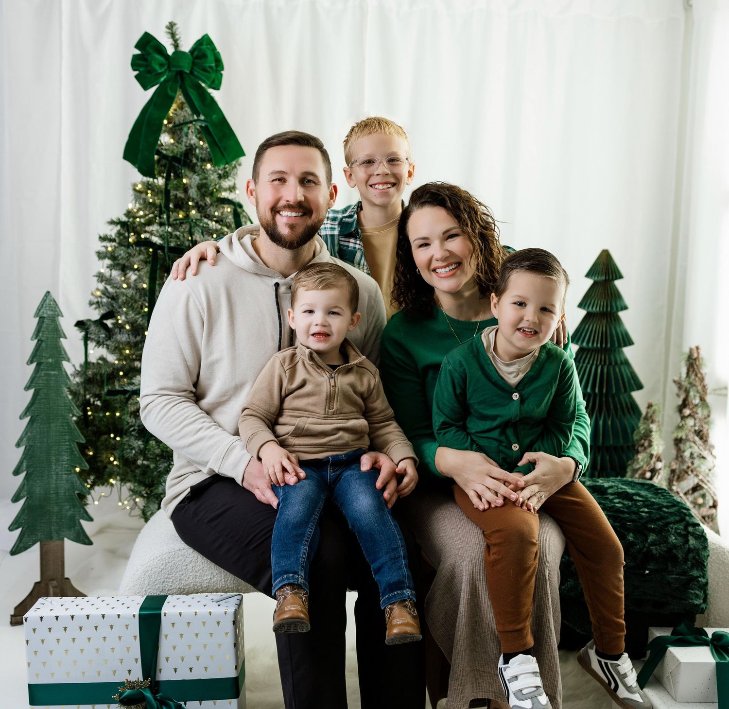 Family wearing coordinated green and brown outfits sitting on white bench with holiday decorations in background.