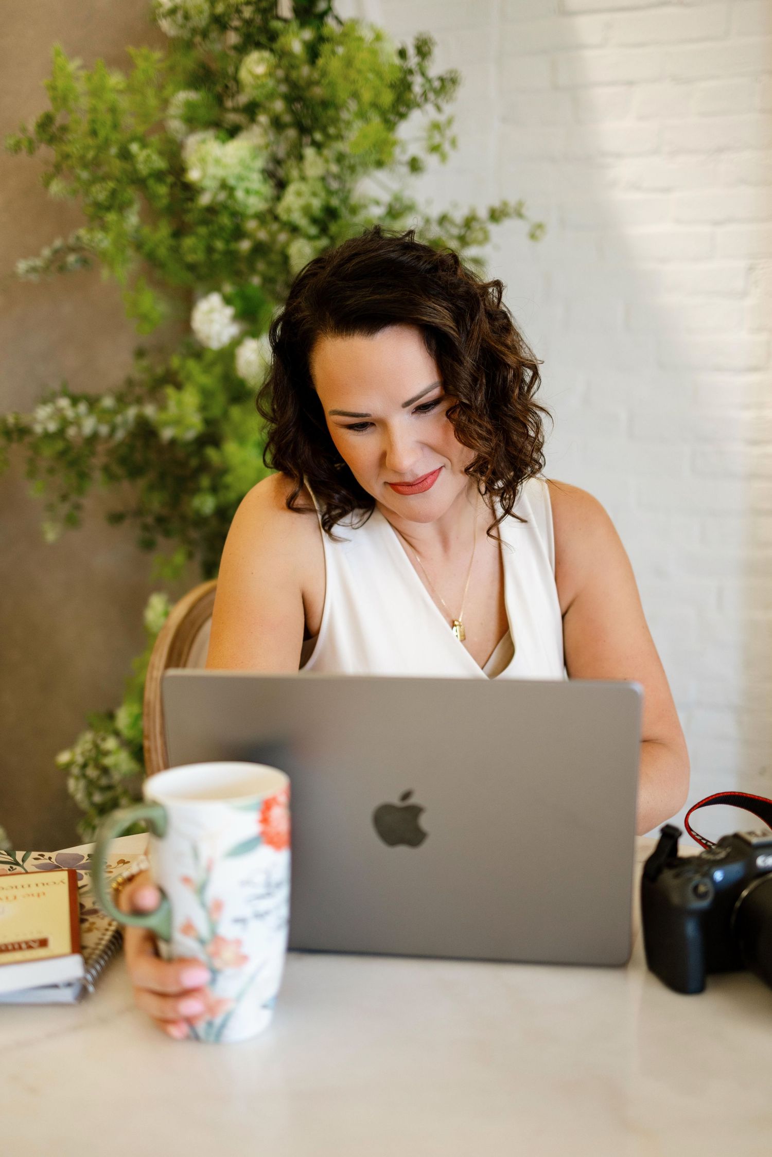 A person with dark curly hair works on a MacBook laptop at a desk with a coffee mug and green plant in the background.