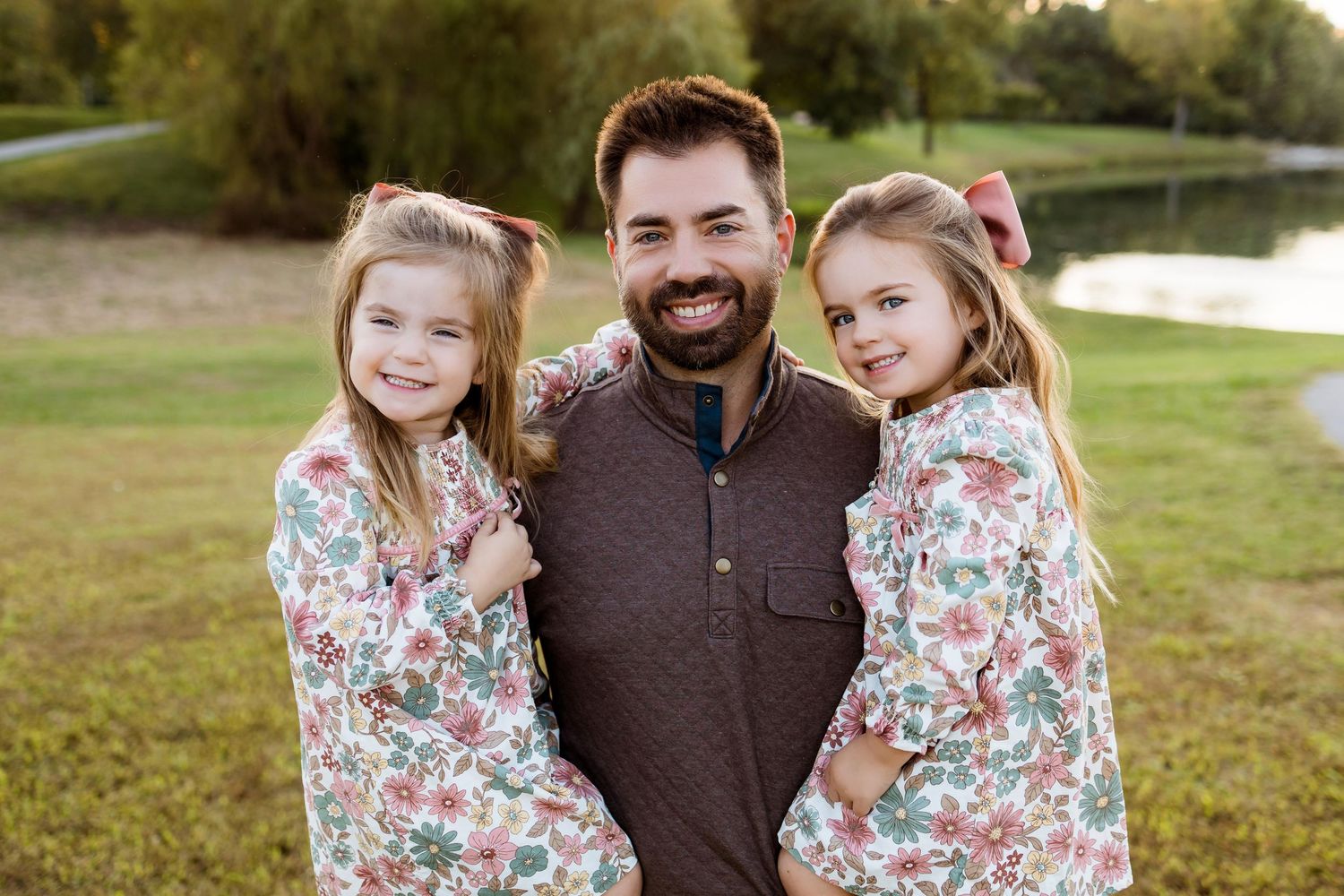 A family poses together outdoors wearing coordinated outfits with floral dresses and a brown sweater near a pond.