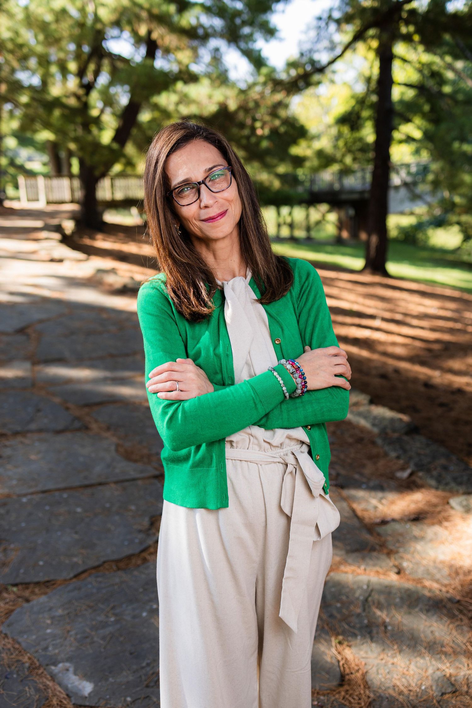 A person in a bright green cardigan and beige dress poses outdoors in a sunlit park setting with trees in the background.