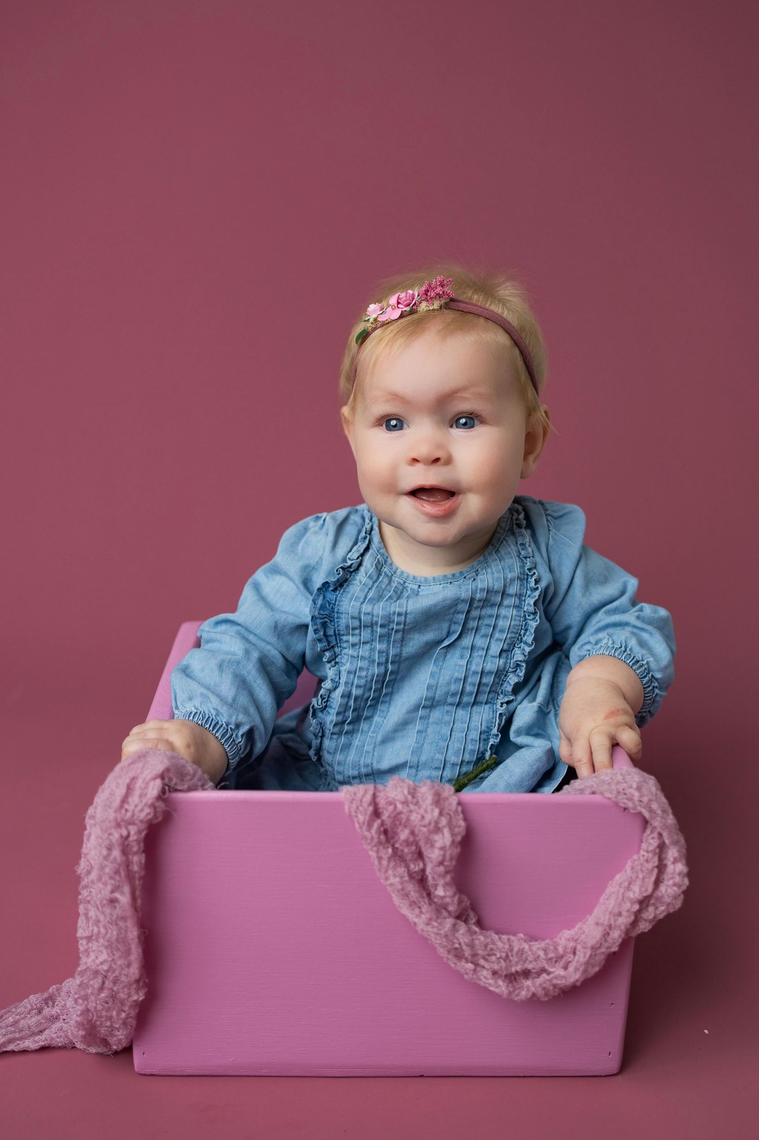 Baby wearing denim dress sits in pink box with lace trim against mauve backdrop in professional photo series.