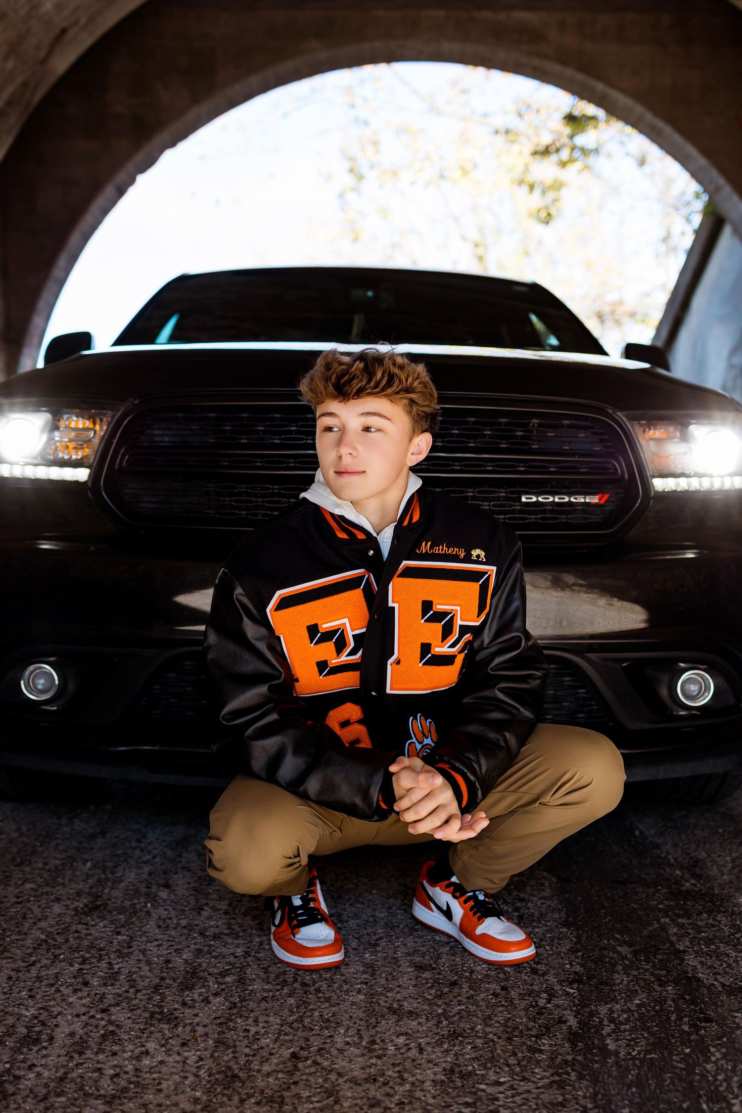 Person in letterman jacket crouches under an archway showing off orange and white sneakers.