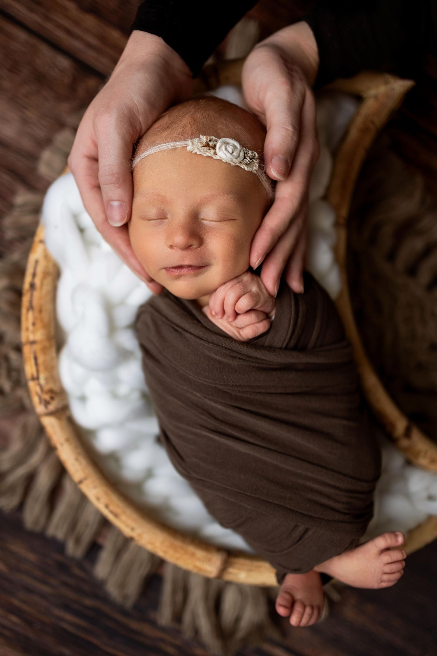 A sleeping newborn swaddled in brown fabric rests peacefully in a basket filled with white fluff.