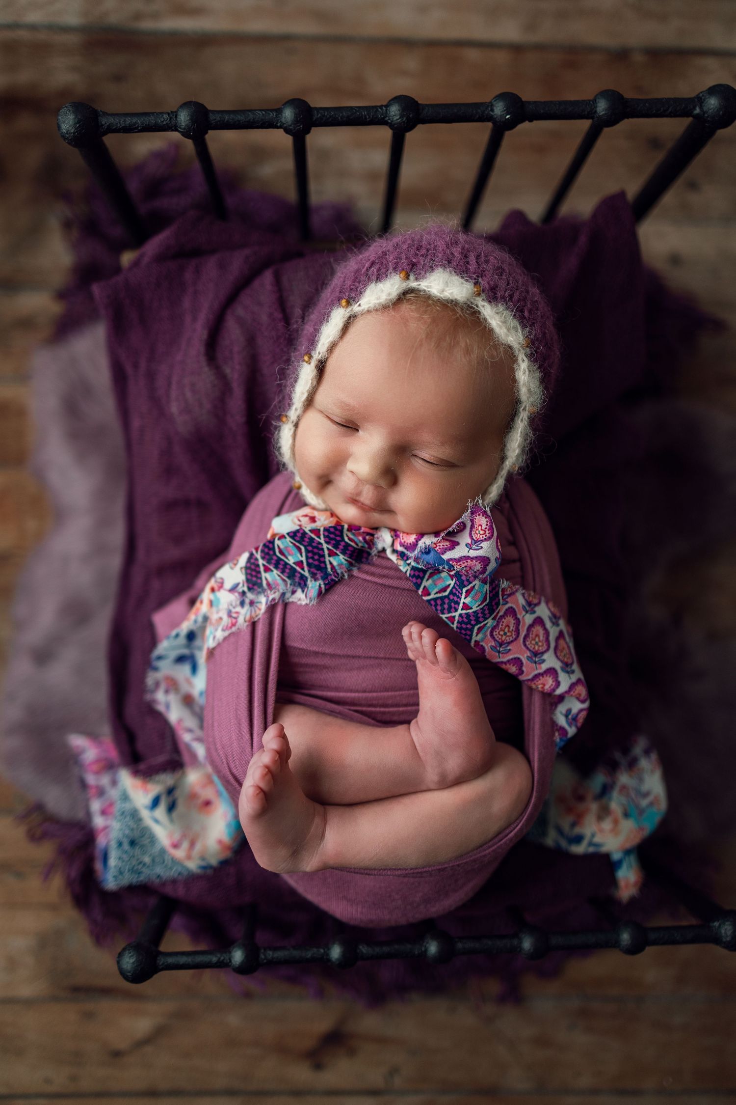 A collection of newborn photography poses on a rustic wooden surface with purple wraps and floral accessories.