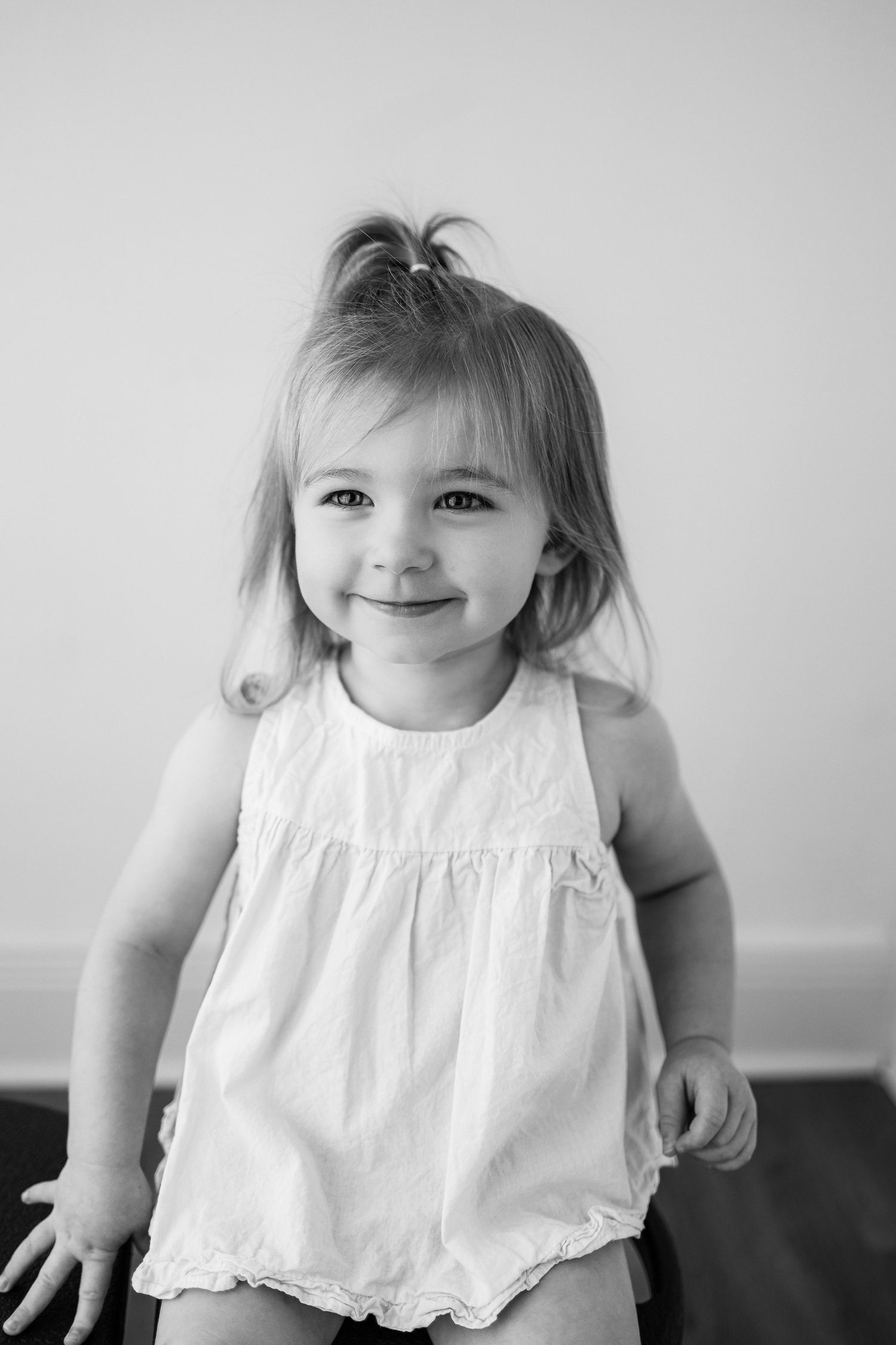 Black and white portrait of a cheerful young child in a white ruffled dress posing against a plain backdrop.