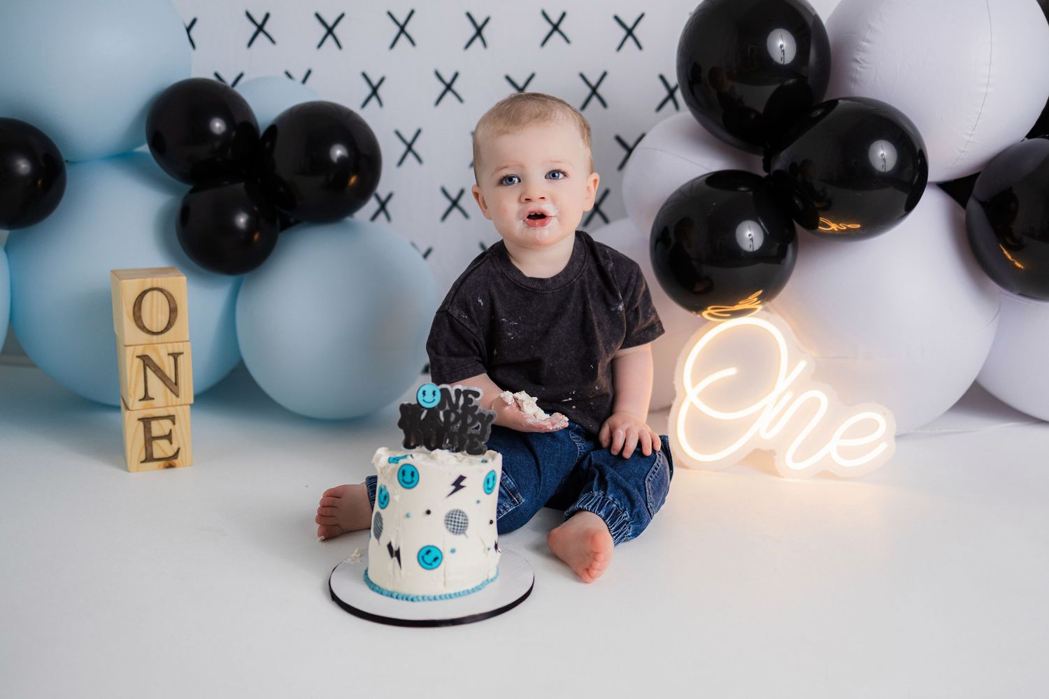 First birthday cake smash photo with black and blue balloons, wooden blocks spelling ONE, and a neon light sign decoration.