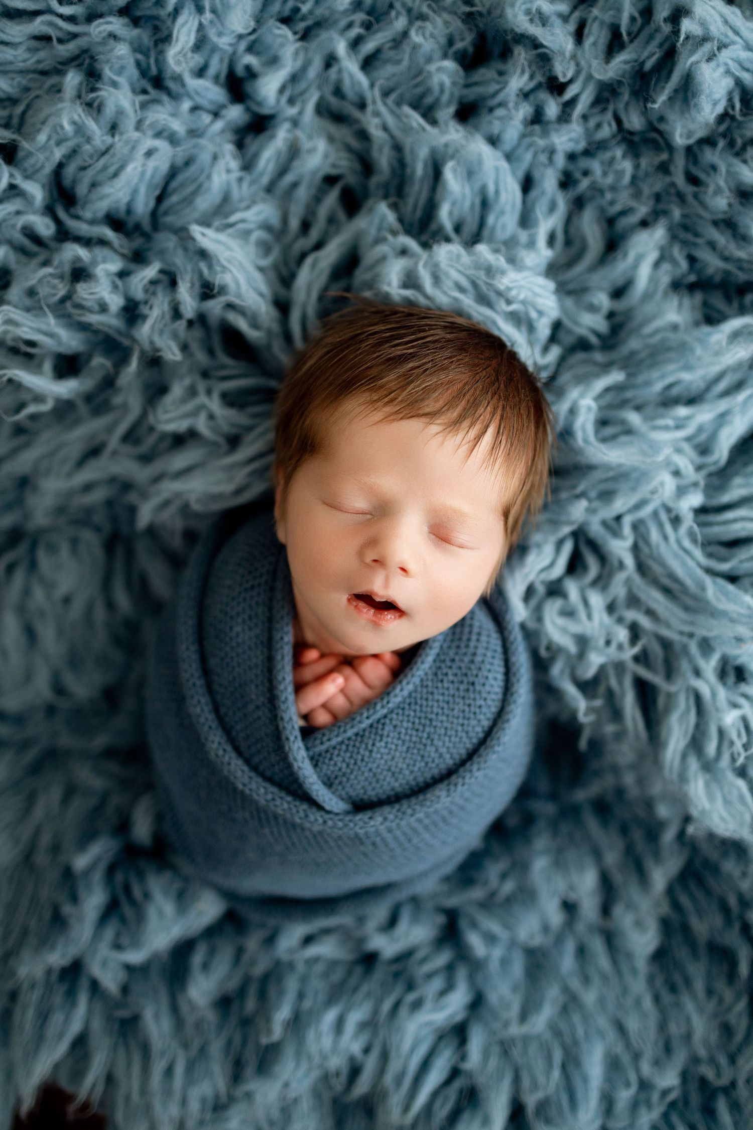 Sleeping newborn baby wrapped in gray knit blanket on fluffy blue fur background.