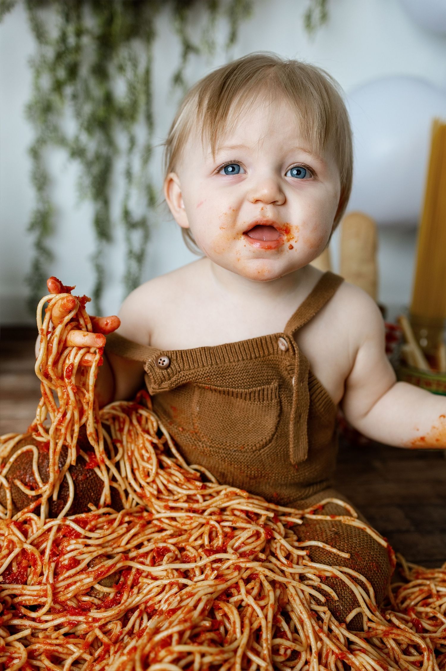 A baby sits in a pile of spaghetti noodles wearing brown overalls while making a playful mess during mealtime.