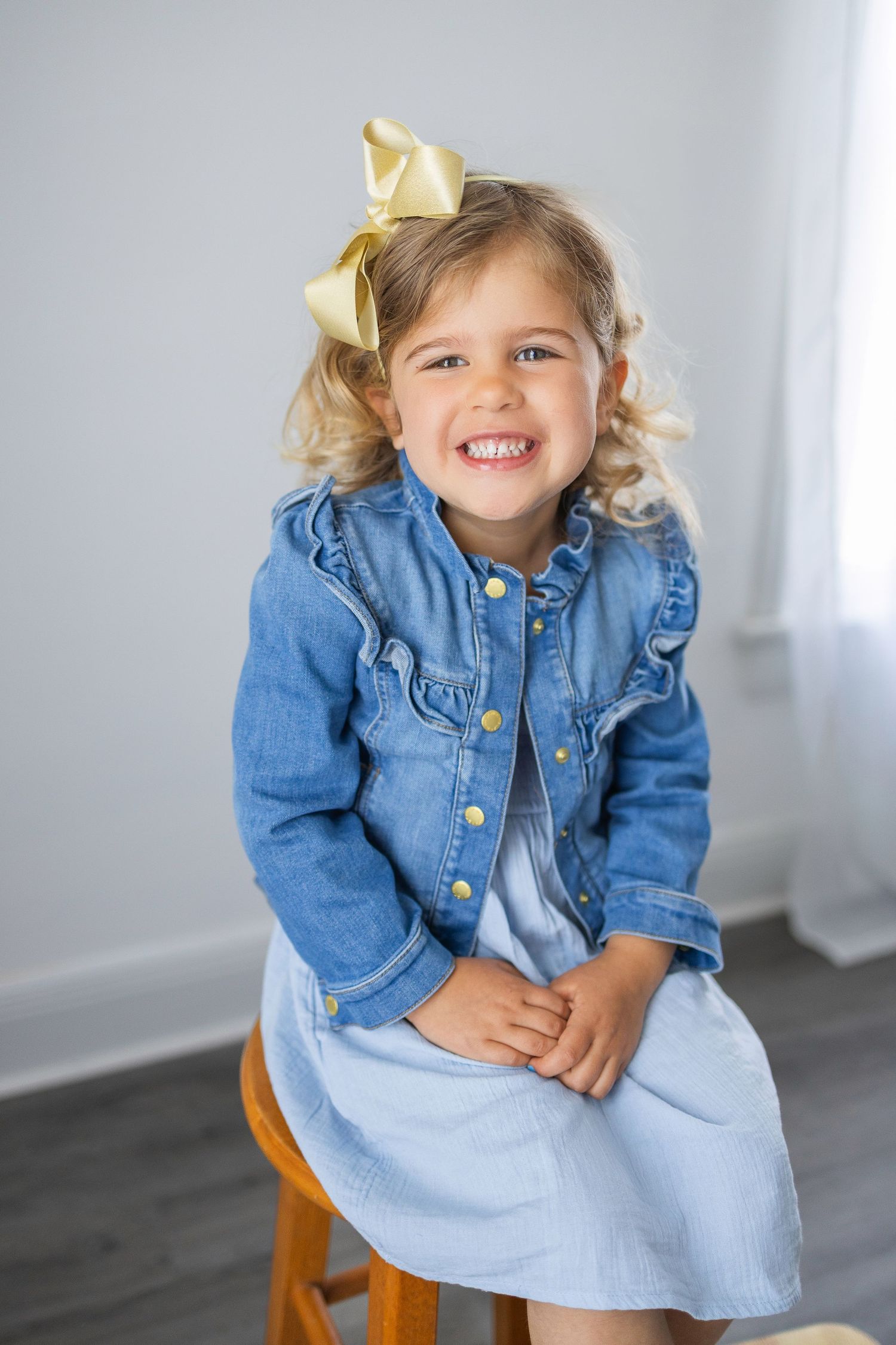 Young child in denim jacket and yellow bow sits smiling on wooden stool in bright room.