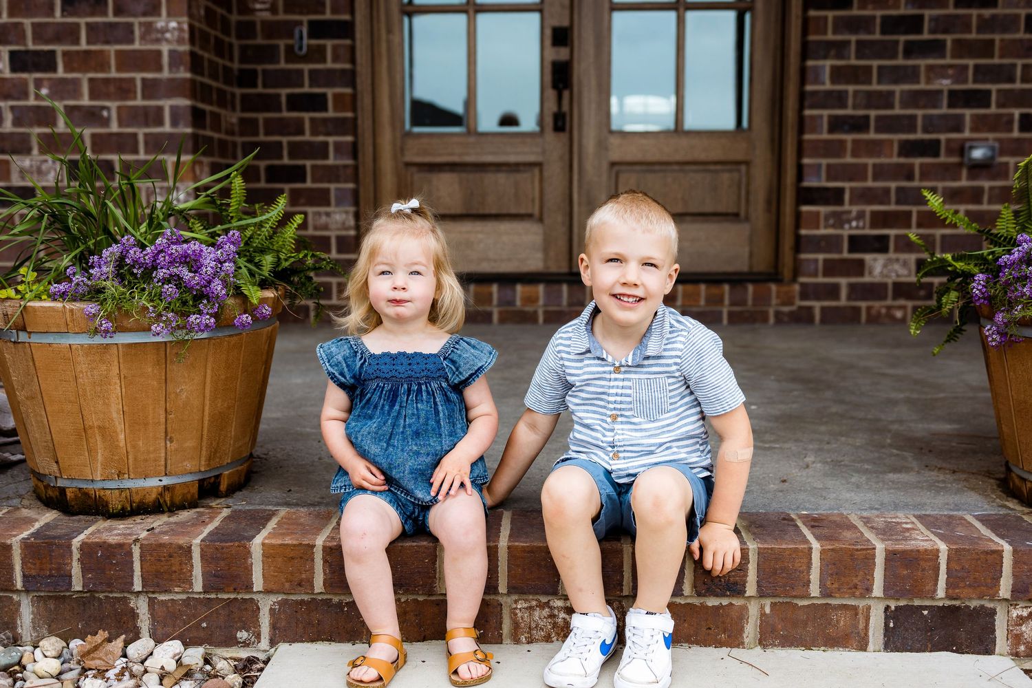 Two children sitting together on brick steps with wooden planters of purple flowers.