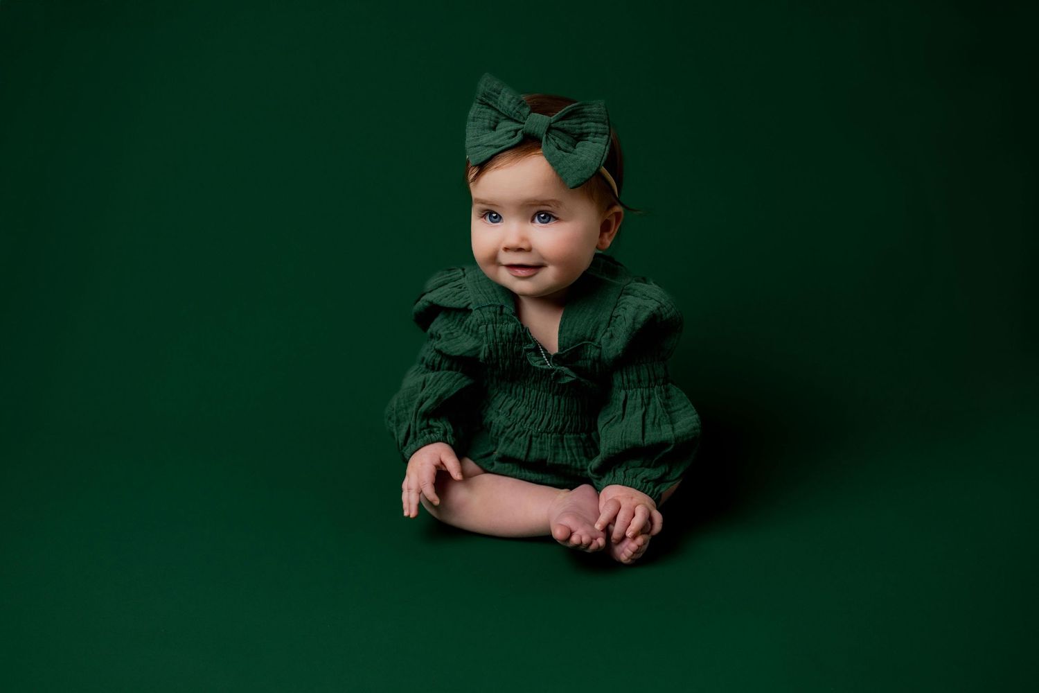 Baby in dark green velvet dress poses against emerald background in holiday portrait series.