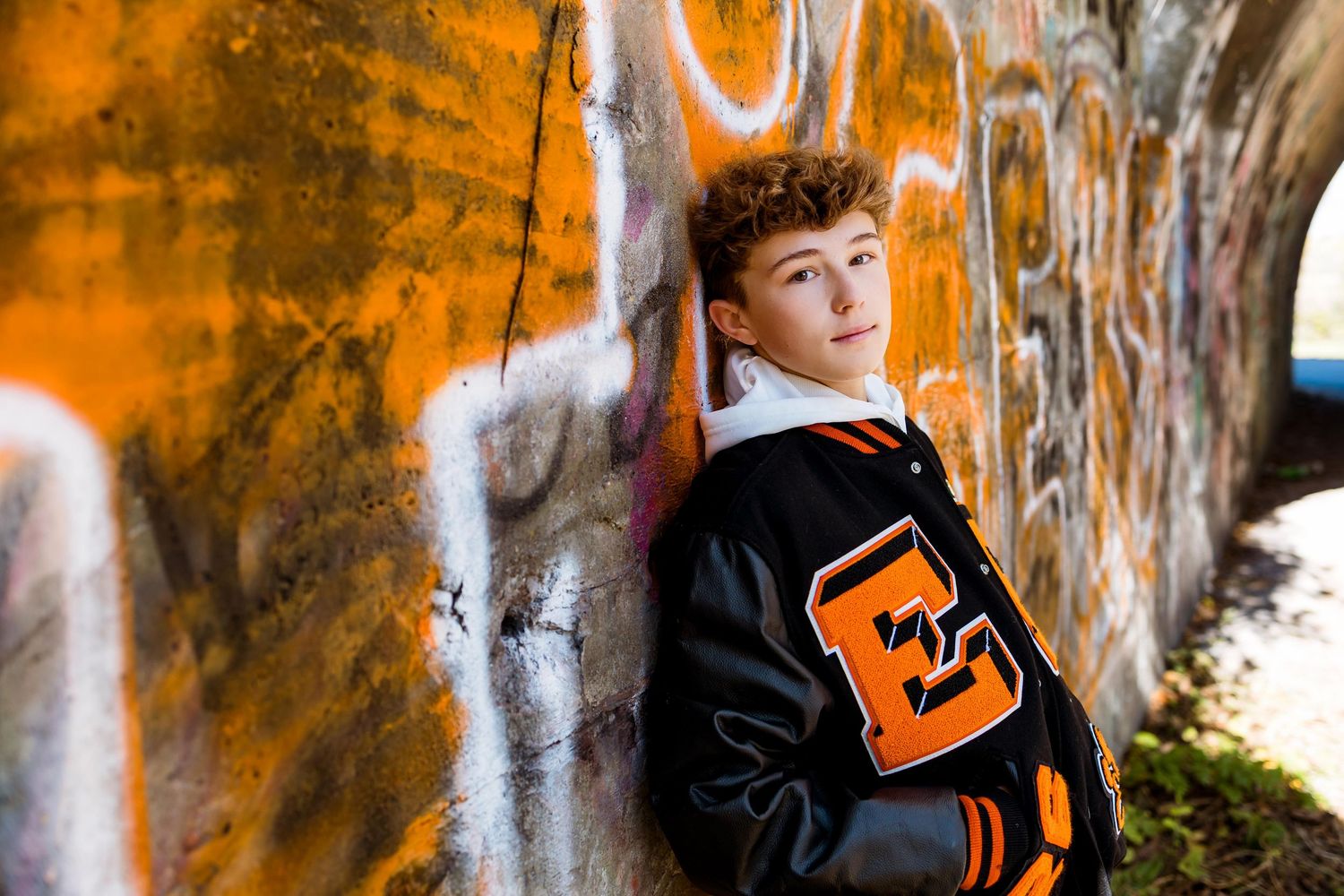 A person in a black jacket poses against a graffiti wall with orange and white street art.