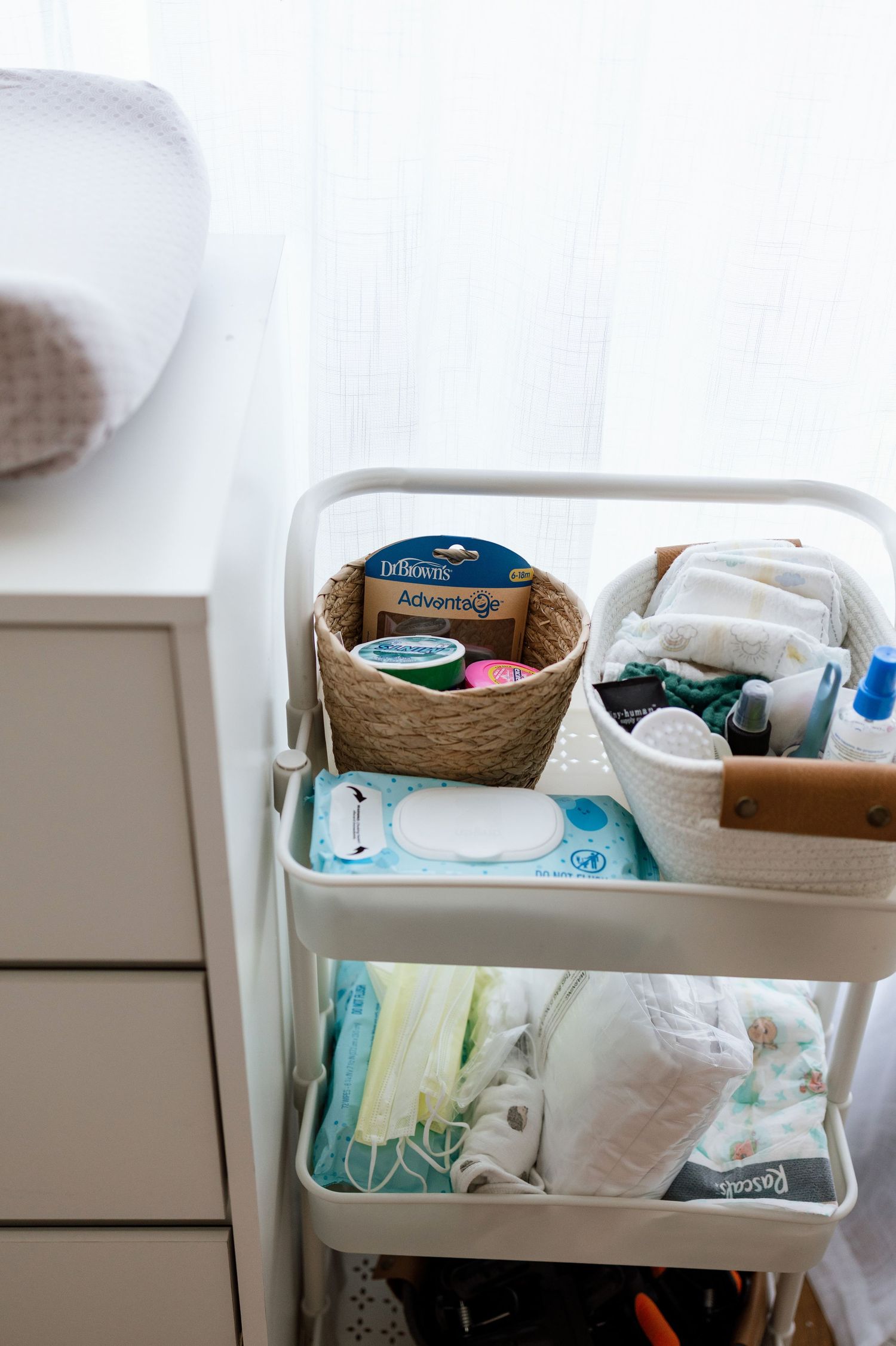 White rolling cart organizer with baby essentials, diapers, wipes, and toiletries in wicker baskets for nursery storage.