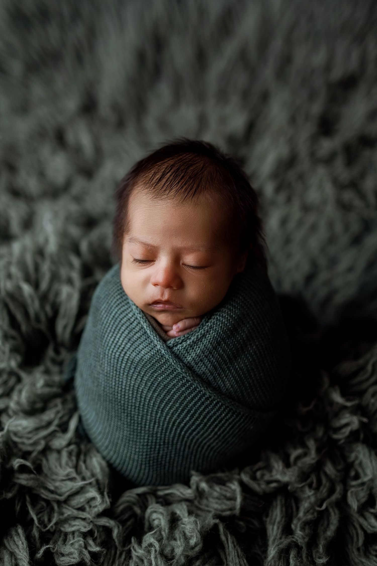 Peaceful newborn photography series showing baby wrapped in dark teal swaddle against textured gray backdrop.