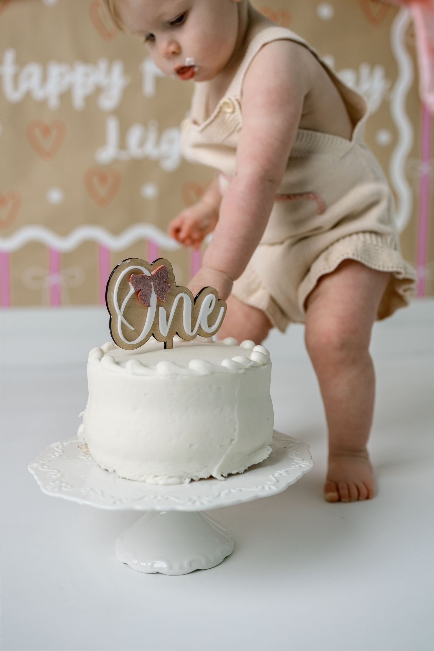 Close-up shots of tiny feet reaching for a white birthday cake on a cake stand against a decorated backdrop.
