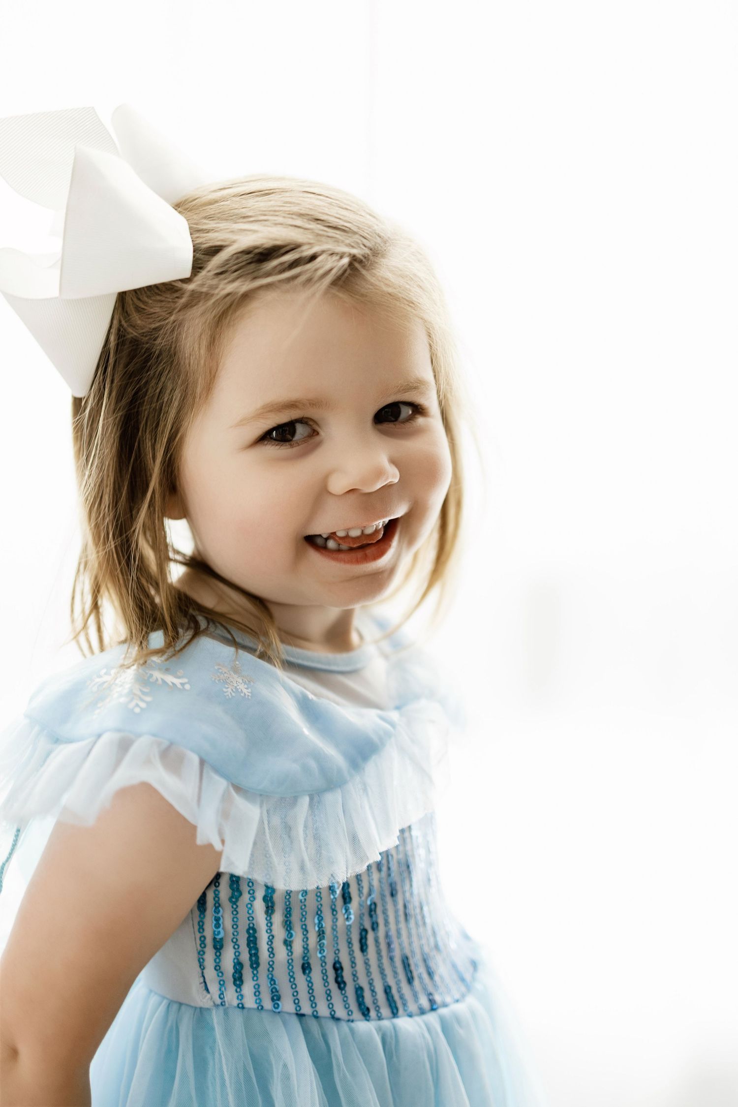 Child wearing a light blue ruffled dress smiles in a bright white studio setting.