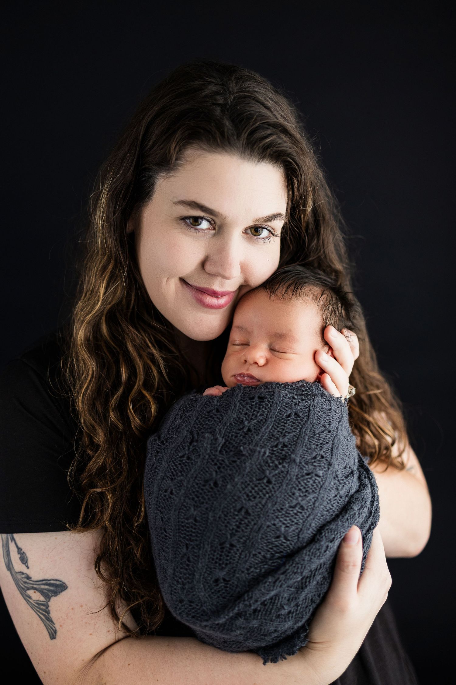 Beautiful newborn portrait showing a sleeping baby swaddled in dark gray wrap against black background with parent.