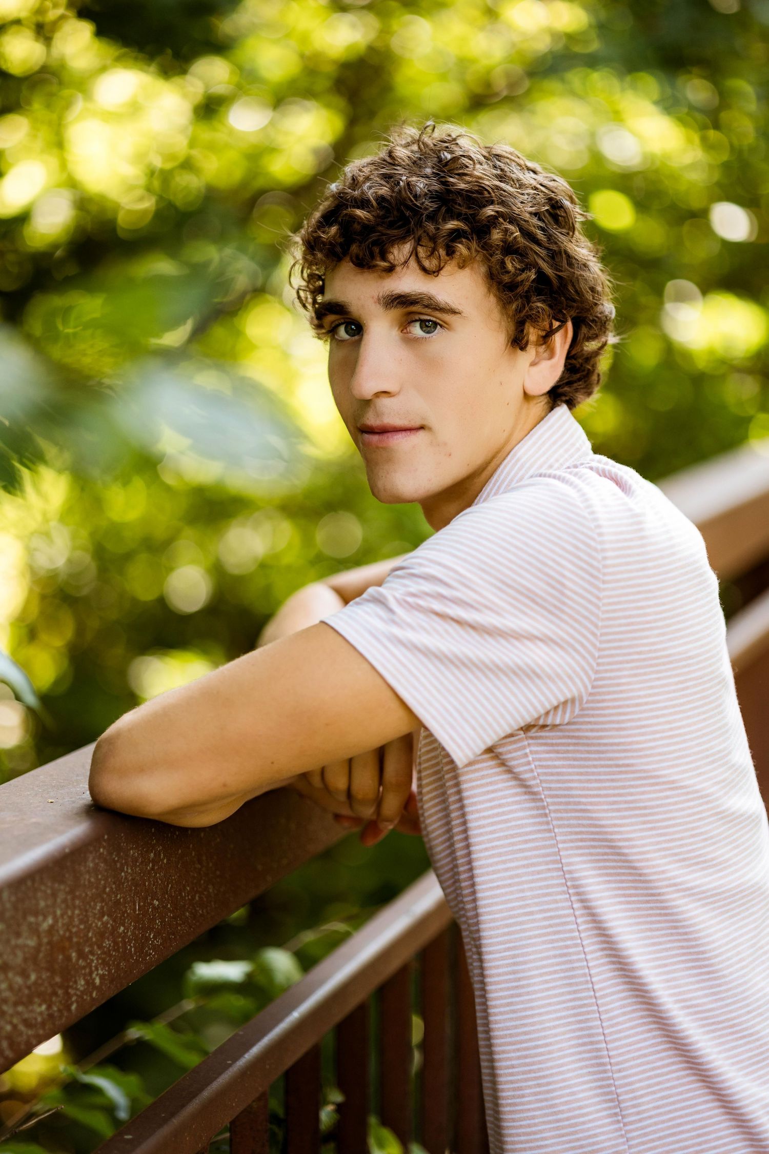 A young person in a white button-down shirt leans casually against a wooden railing in a sunny outdoor setting.