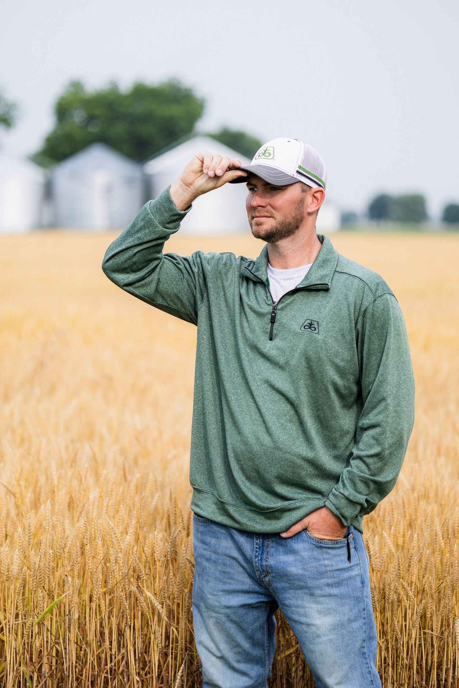 Person wearing a green sweater and baseball cap stands in a golden wheat field while looking into the distance.