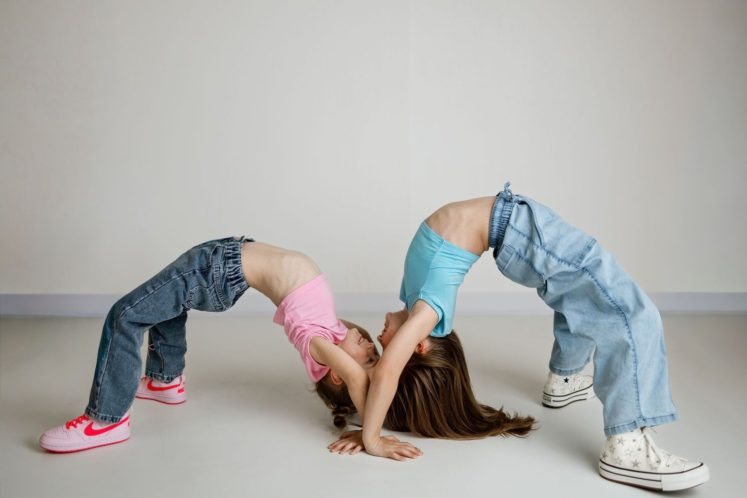 Two girls performing backbend bridge poses facing each other in jeans and colorful tops on a light gray background.