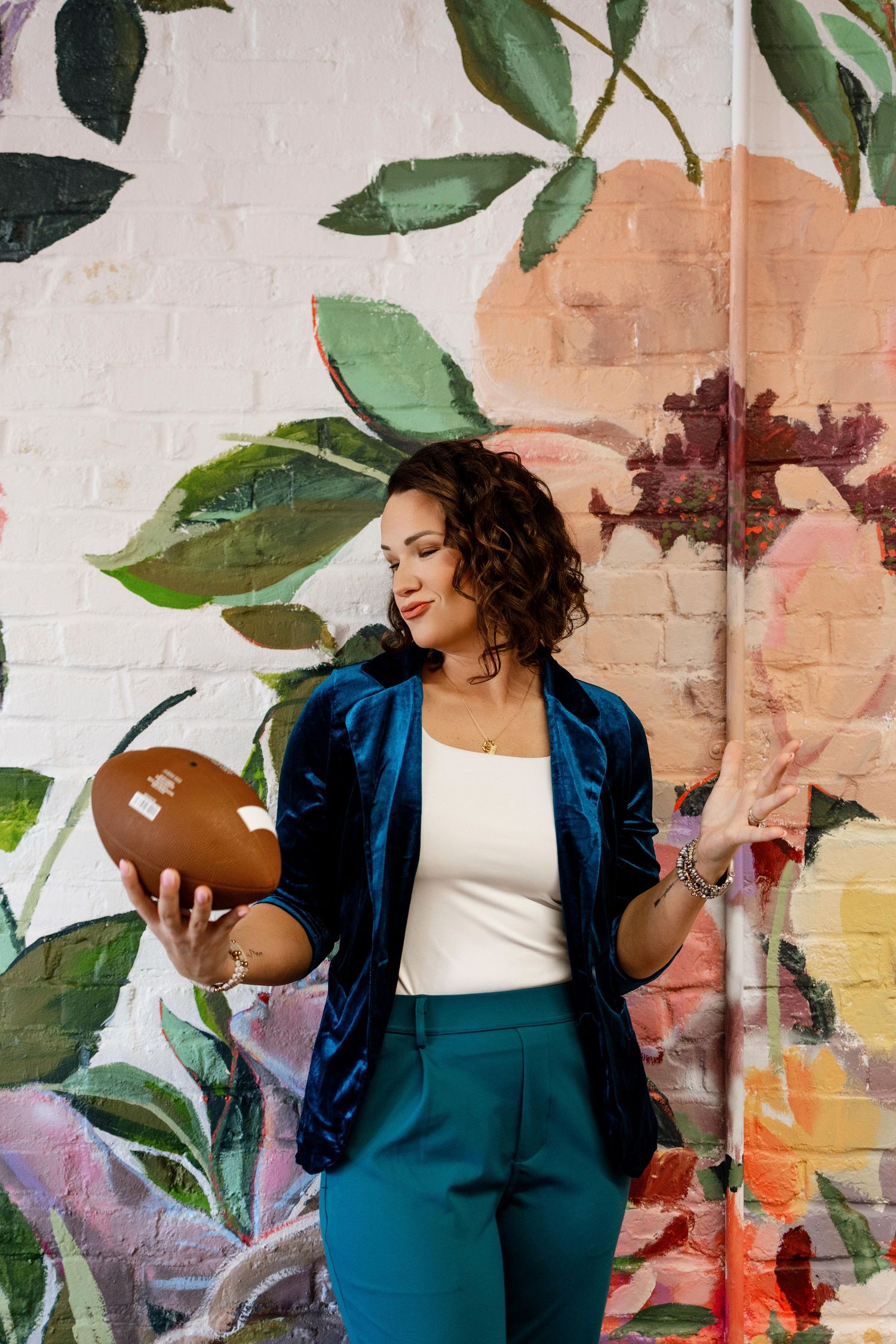 Photographer in blue velvet blazer poses against artistic wall with green plants.