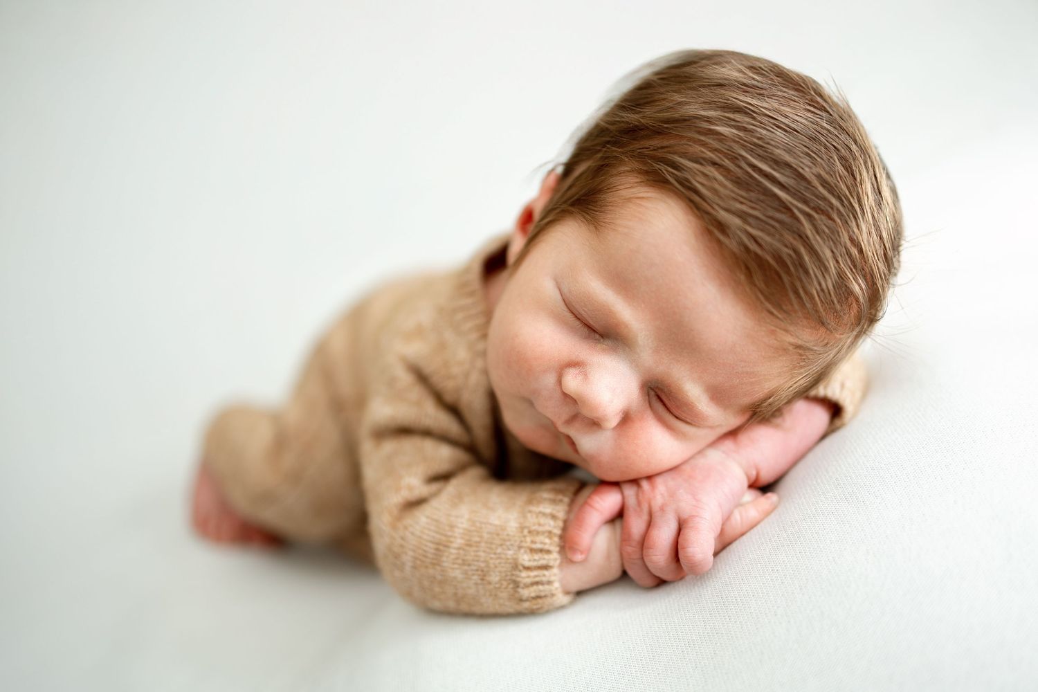 Sleeping newborn baby in beige sweater lying on white background during professional photo session.