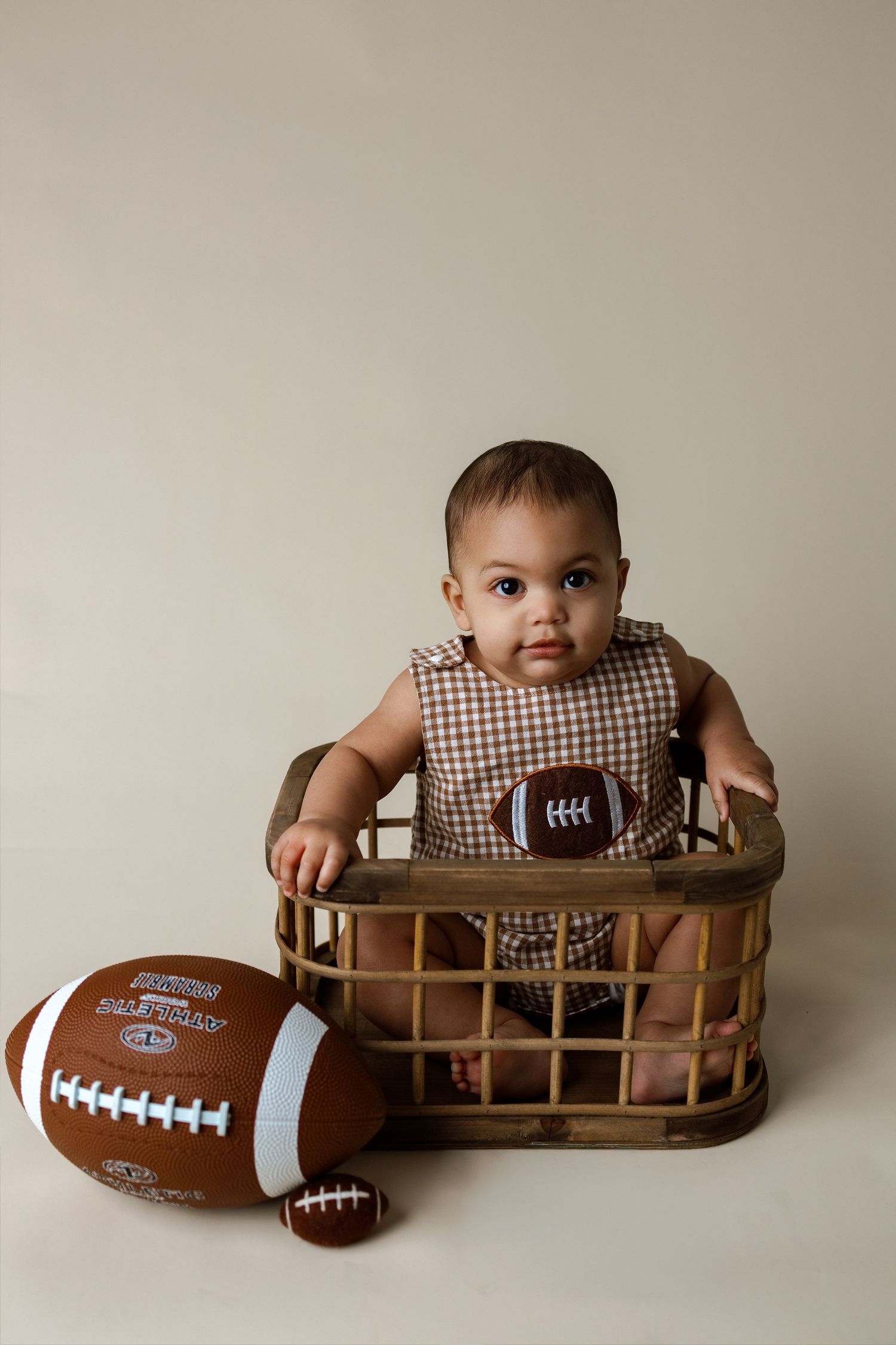 A baby in a checkered outfit poses with a football on a wooden bench during a studio photoshoot.