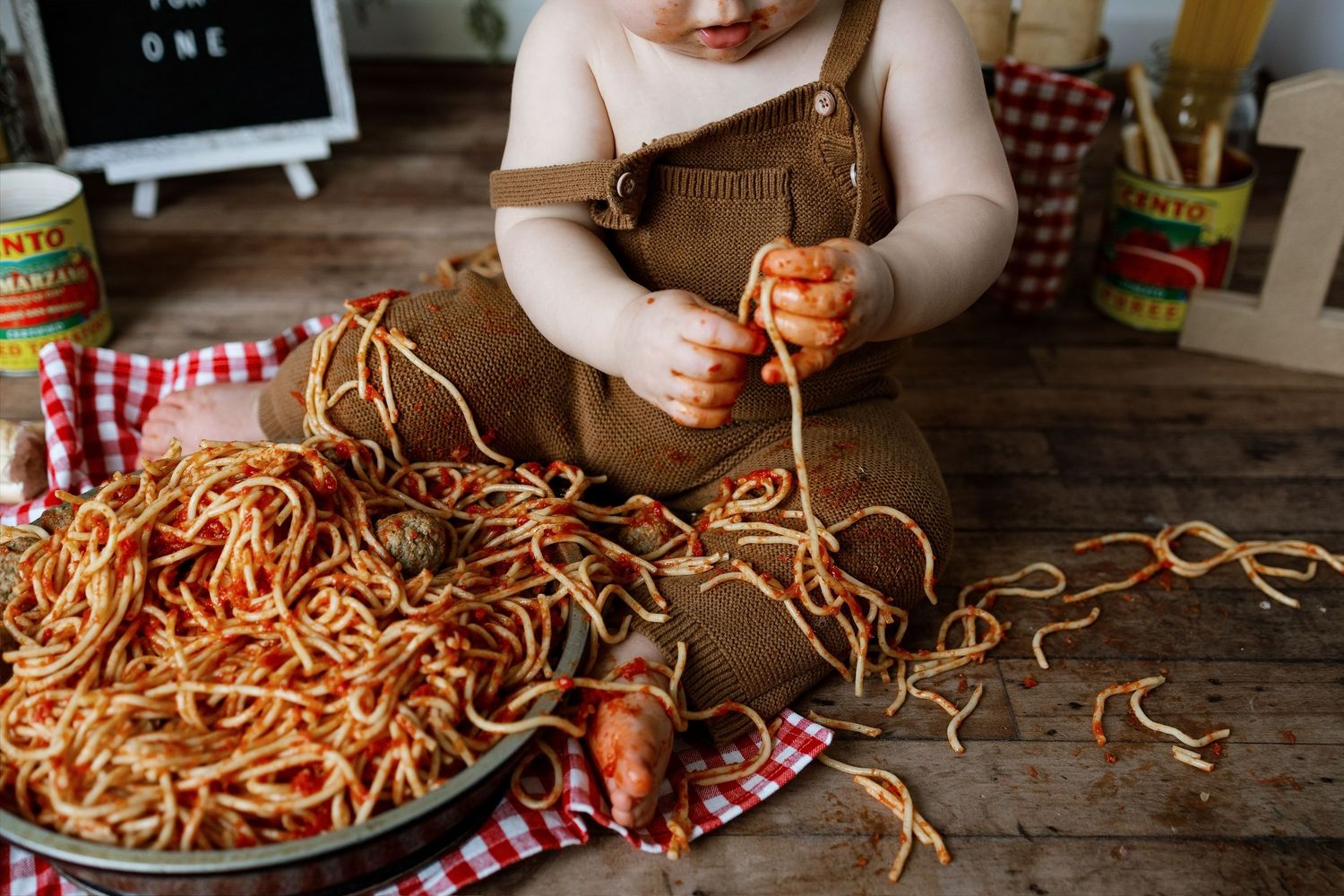 A messy plate of spaghetti with tomato sauce sits on a checkered cloth while small hands hold noodles at a wooden table.