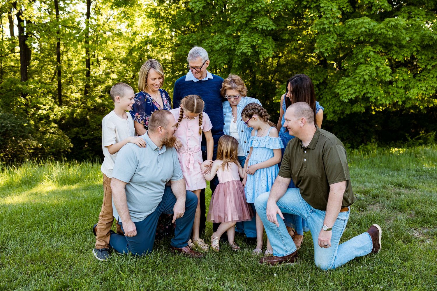 Multi-generational family gathering outdoors in a sunlit garden setting with green trees in background.
