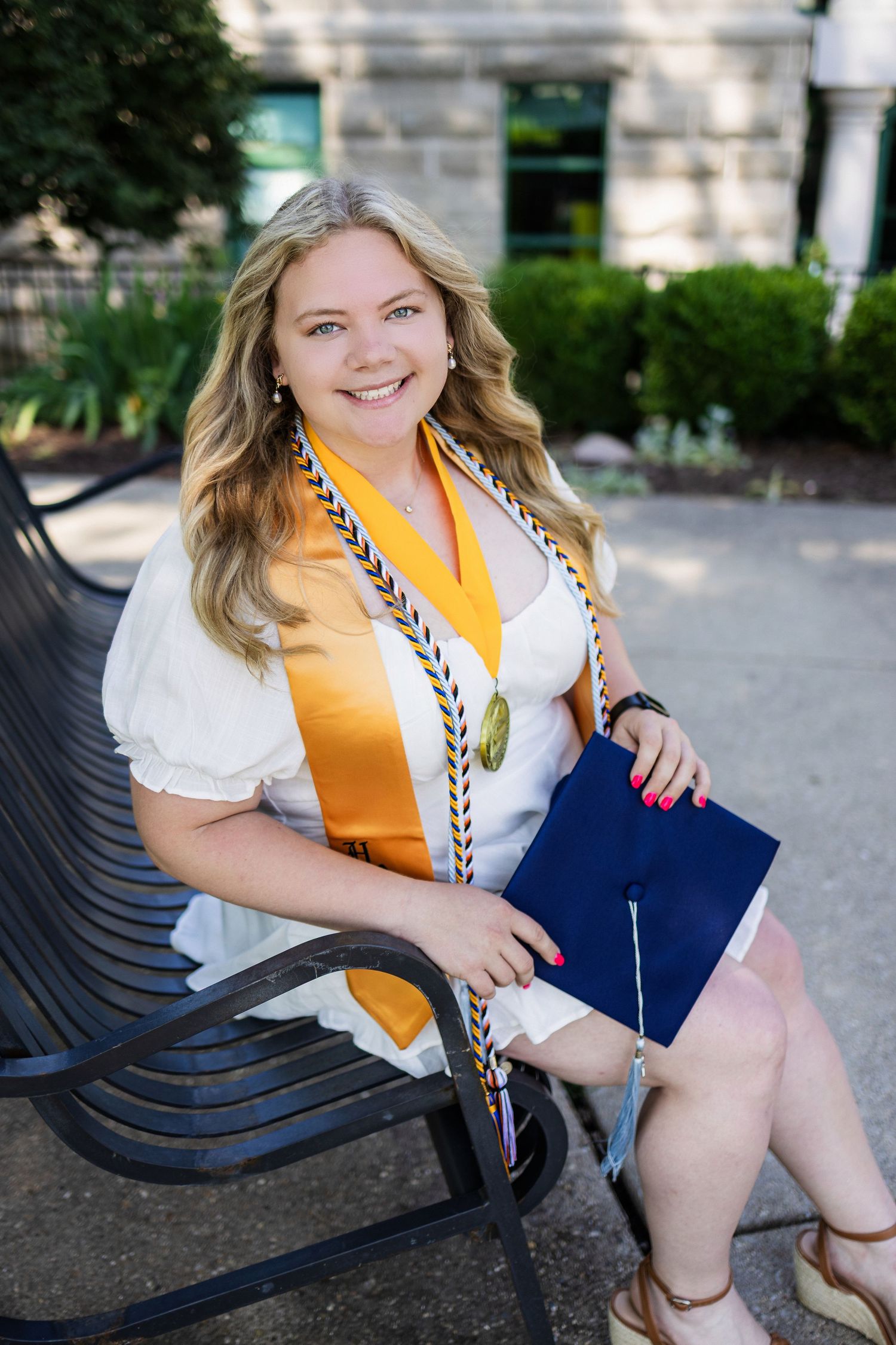 Graduate in white blouse and gold sash sits on bench holding diploma while posing for outdoor graduation photo.